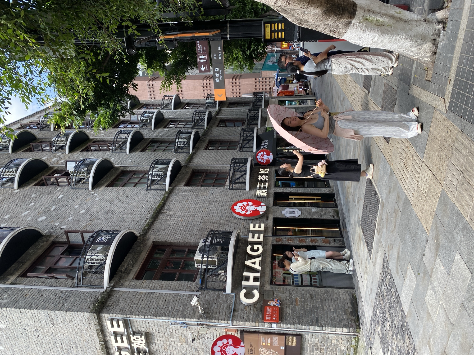 The image depicts a street scene in Yuzhong District, China. Two women are standing on a sidewalk next to a brick building. The woman on the left is holding a pink umbrella and looking down. She is wearing a gray tank top and a white and gray striped skirt. The woman on the right is wearing a light gray t-shirt and white patterned pants. She is holding a phone and appears to be looking at it. A third person, partially visible, is standing behind them. The building behind them has multiple floors with arched windows and balconies. There are several signs visible, including one that reads "CHAGEE" and another that says "霸王茶姬" which translates to "Ba Wang Cha Ji" or "Overlord Tea Princess," a popular Chinese tea chain. Other signs indicate "Public Restroom" and "Daijia Alley." The scene appears to be taking place during the day, with sunlight casting shadows. The overall atmosphere seems relaxed and casual, with people strolling along the street.