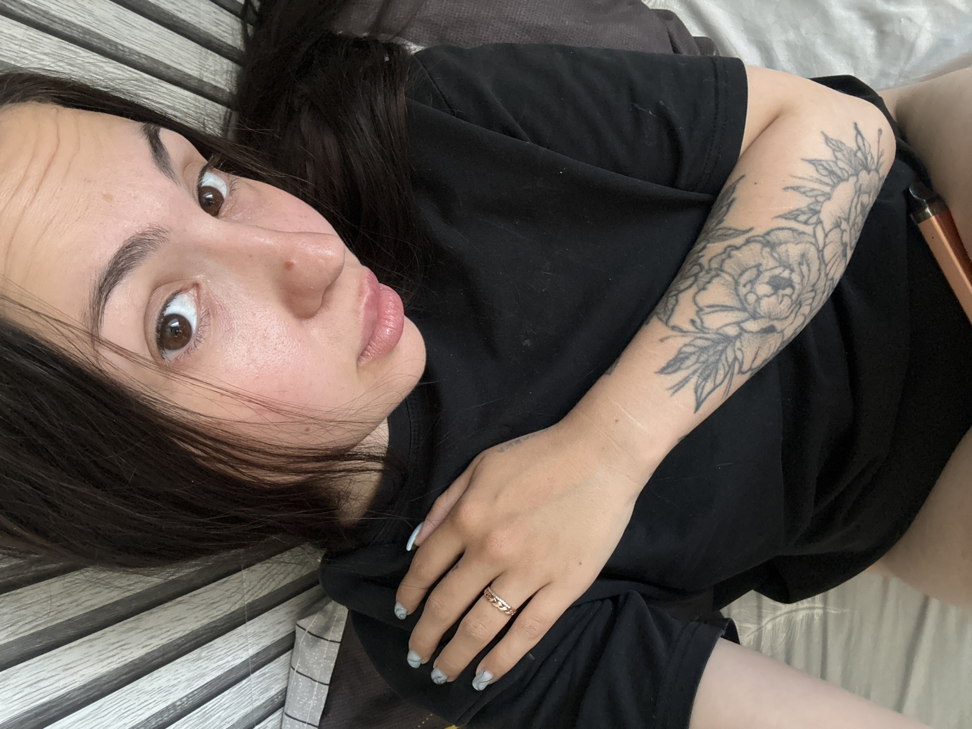 A young woman with long dark hair and brown eyes lies on a bed. She is wearing a black t-shirt. Her left arm is resting on her chest, and she has a ring on her left ring finger and a tattoo of flowers on her left forearm. The background appears to be a headboard with vertical slats. The scene is indoors, likely a bedroom, with no specific identifiable city or Russian context present in the image.