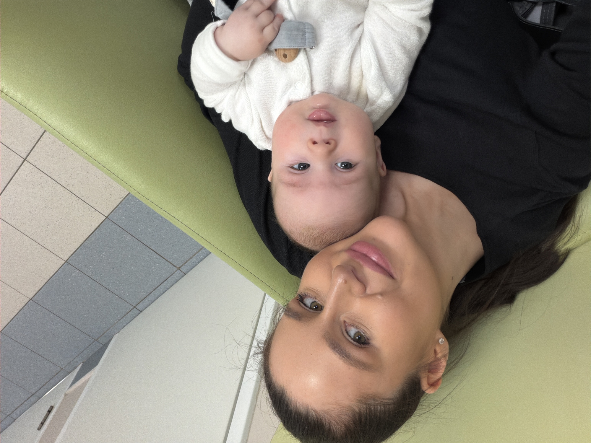 A woman and a baby are lying on a green surface. The woman has fair skin, dark hair pulled back, and is wearing a black top. The baby is wearing a white outfit. They appear to be in a room with gray and white tiled floors. The location cannot be determined as Moscow, Russia from the image alone.