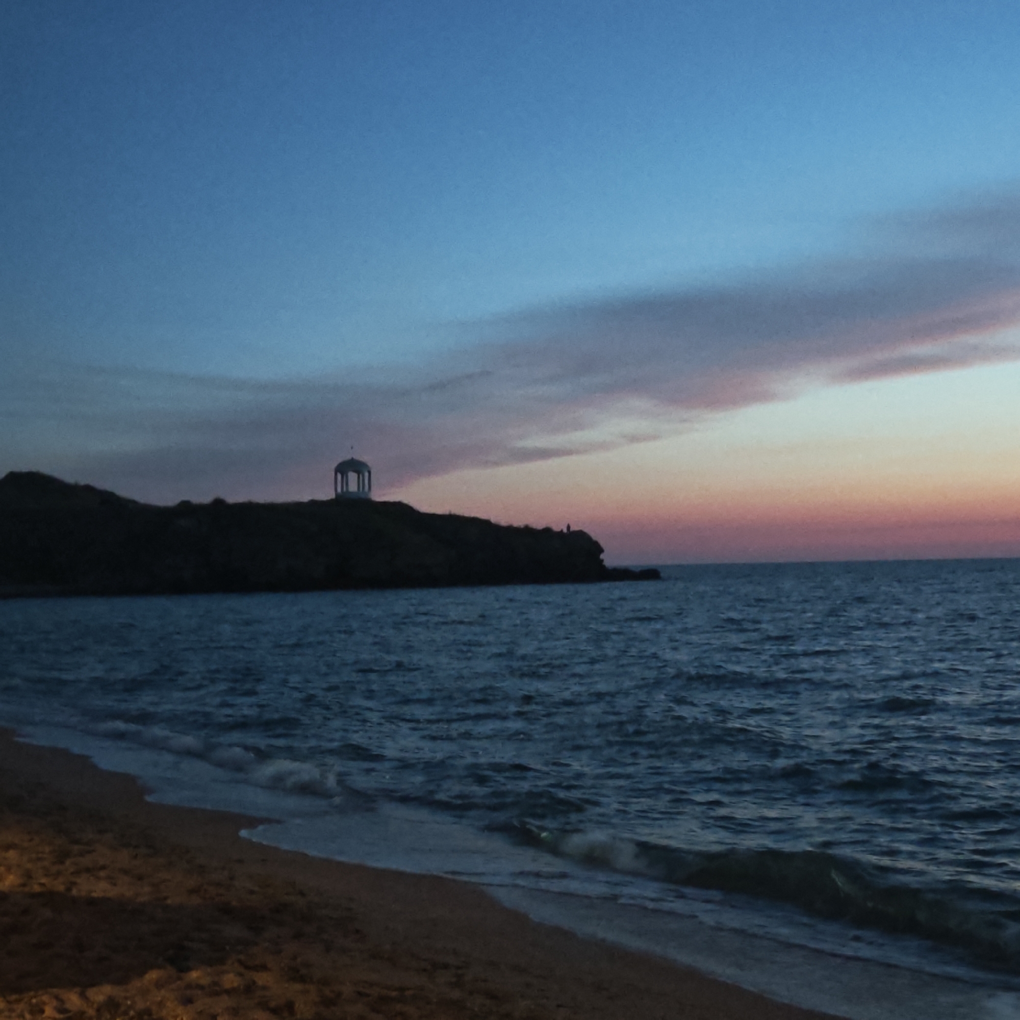 The image depicts a twilight scene at the seashore. In the foreground, a sandy beach stretches along the bottom of the frame. Gentle waves with white foam lap at the shore. The sea itself is a deep blue, with visible ripples on its surface.
In the mid-ground, a dark, silhouetted headland juts out into the sea. Perched atop the highest point of this headland is a white rotunda, a circular structure with a domed roof supported by columns. The rotunda is clearly visible against the darkening sky. There appear to be two small, indistinct figures standing on the edge of the headland, near the rotunda.
The sky above is a gradient of colors characteristic of dusk. The upper portion is a deep, clear blue, transitioning to softer blues and then to hues of pink and orange closer to the horizon. A thin, wispy cloud band is visible across the sky, catching the fading light. The overall impression is one of tranquility and natural beauty at the close of day. The scene is consistent with the coastal landscape found in the vicinity of городское поселение Щелкино, Ukraine, which features such scenic overlooks and seaside vistas.