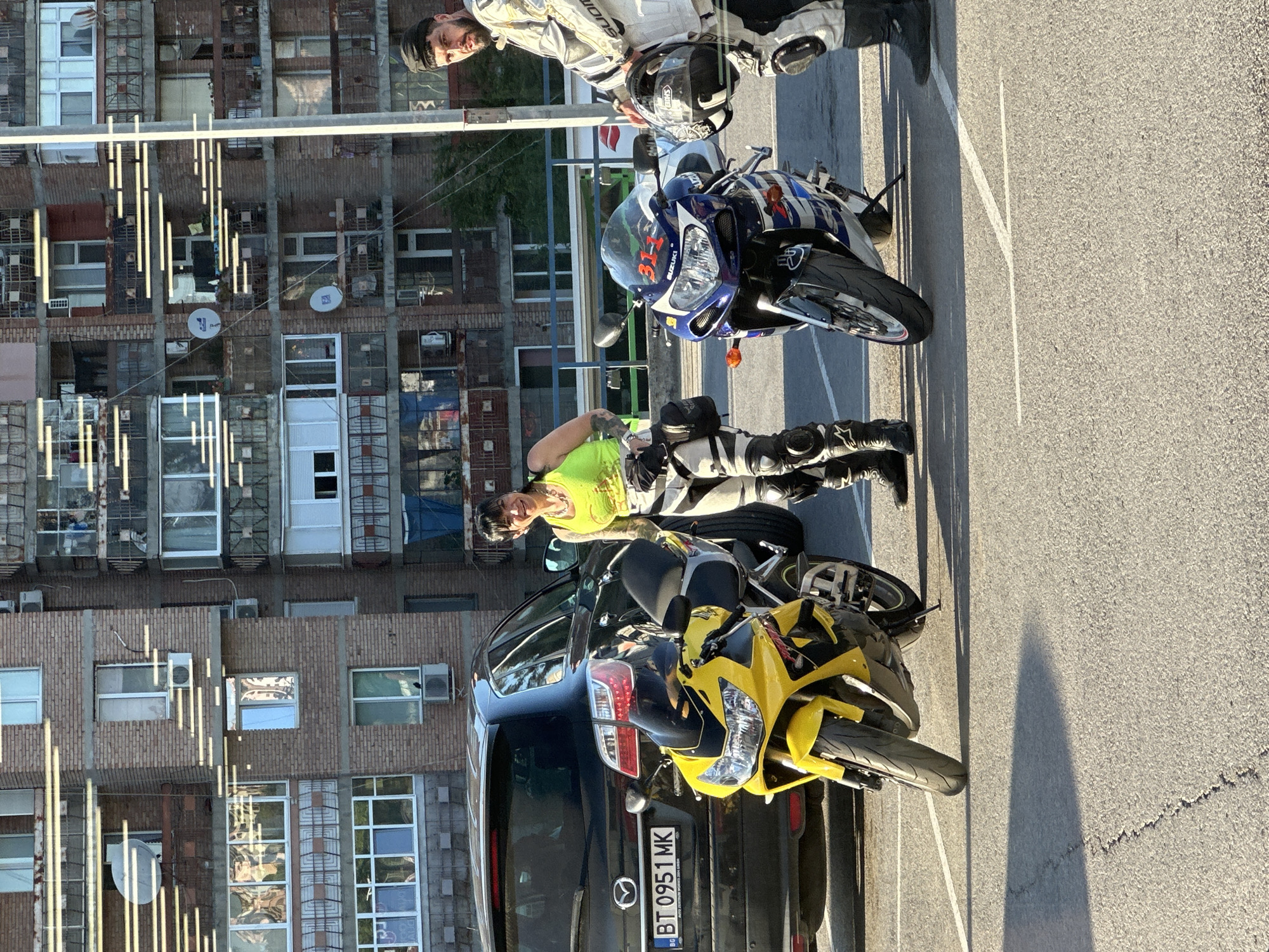 Two people, a woman and a man, are standing next to two motorcycles in a parking lot. The woman is wearing a bright yellow tank top and black and white motorcycle pants. She is smiling and looking towards the camera. The man is wearing a white and black motorcycle suit and a black beanie. He is holding a motorcycle helmet. In the background, there is a dark-colored car and a brick apartment building. The apartment building has many windows and balconies, and a satellite dish is visible on one of them. The scene appears to be outdoors during the daytime, with the sun casting shadows on the ground. The license plate on the car reads "BT 0951 MK". The overall setting suggests a casual encounter or stop during a motorcycle ride. The presence of the license plate format "BT" indicates the location is likely in Bulgaria, and specifically Veliko Tarnovo, given the context provided.