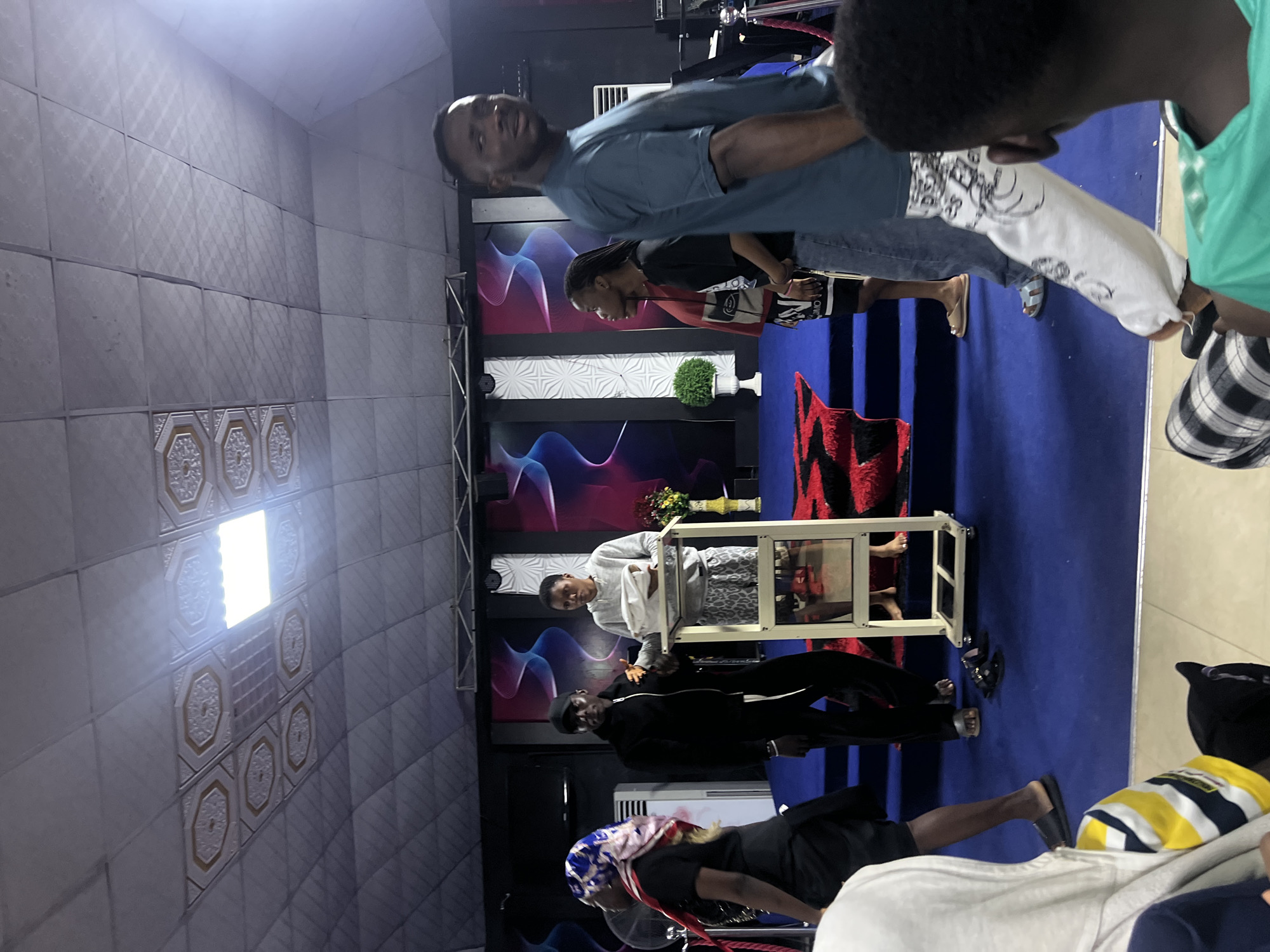 The image shows a group of people gathered in what appears to be an indoor event space, possibly a church or conference hall, located in Nkpor, Nigeria. The setting has a stage area with a blue carpet and red accents. A white podium is present on the stage, and a woman in a gray hoodie stands behind it, holding a white object. Beside her, a man in a black jacket and cap is looking towards the woman. To the left of the stage, a woman with a colorful headscarf is walking away from the camera. In the foreground, a person in a white hoodie is seated, and another person in a teal t-shirt is looking towards the right. The ceiling is made of suspended tiles with decorative elements and fluorescent lighting. A fan is visible to the left of the stage. The overall atmosphere suggests a congregational or performance event.