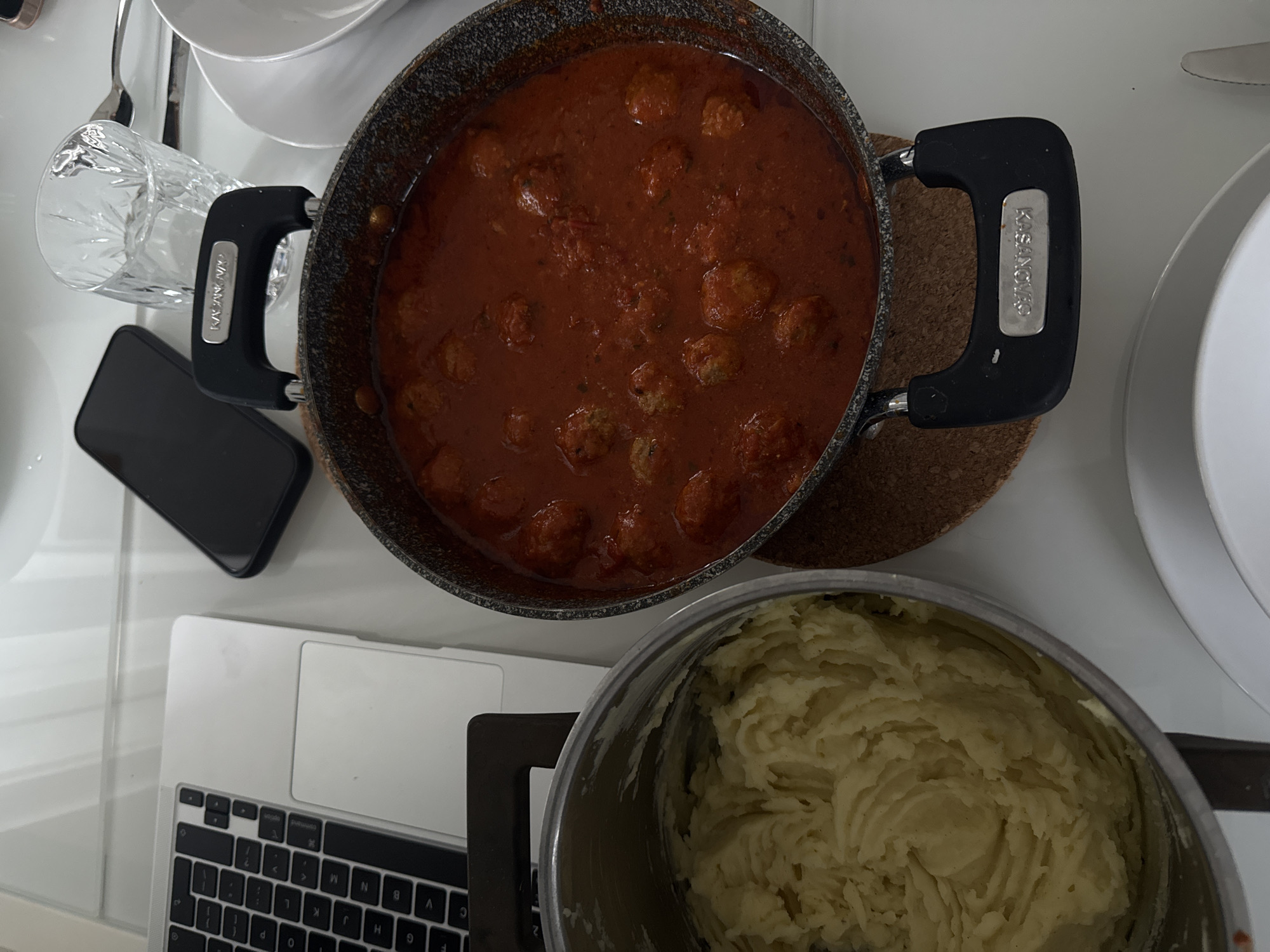The image shows a flat lay on a likely white table featuring several items: a laptop with a visible keyboard, a black smartphone, a clear glass, white plates stacked, a pan filled with meatballs in tomato sauce, and a pot containing mashed potatoes. The pan of meatballs has "Kasanova" printed on its handle. The scene suggests a meal preparation or consumption environment indoors. There's no contextual information confirming it is in Milan, Italy, but based on the presence of the brand "Kasanova," it is plausible the location might be in Italy, as this is an Italian brand. The environment is generally clean and organized.