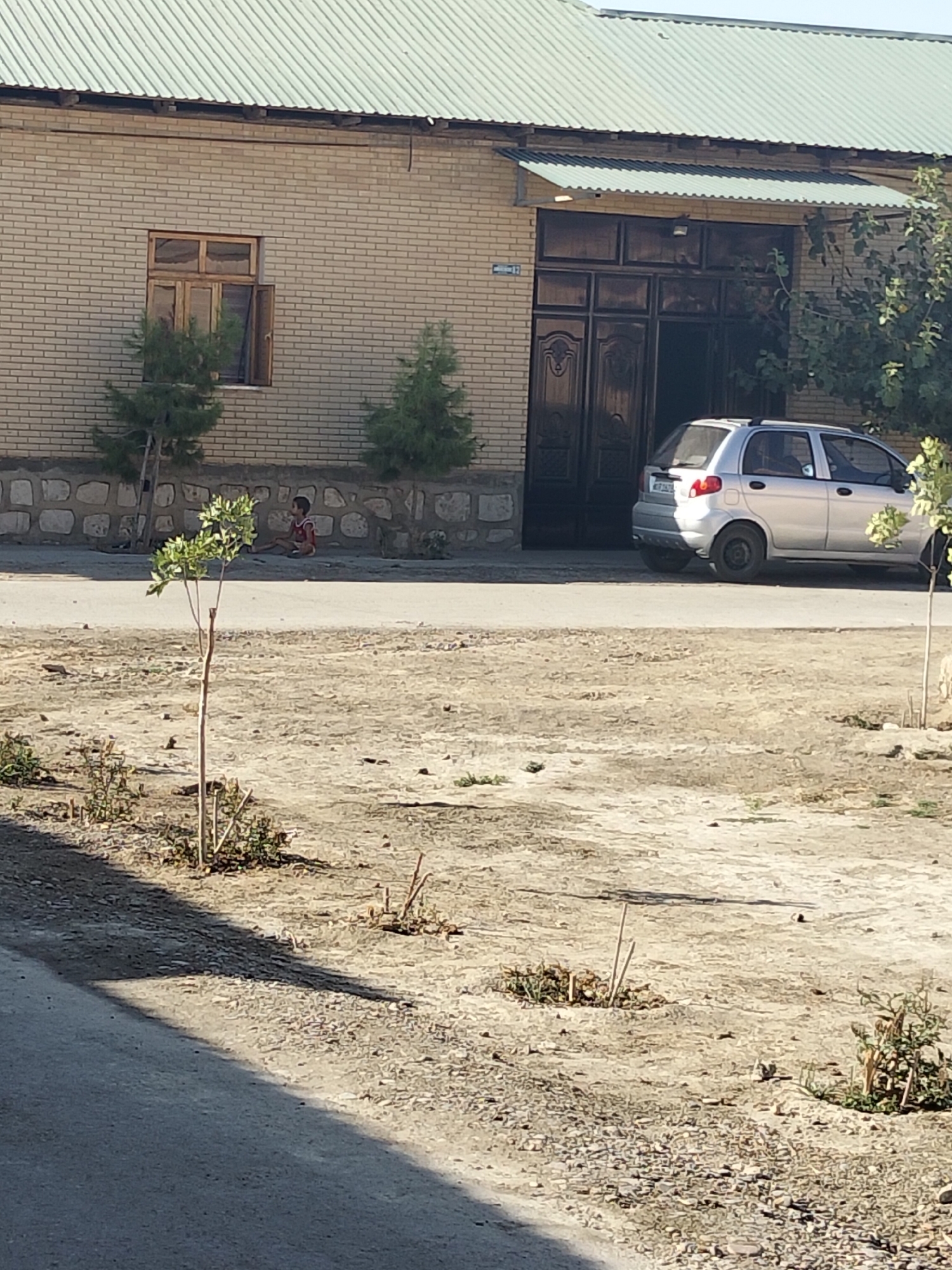 In Kogon City, Uzbekistan, a silver hatchback is parked on a paved street in front of a light-brick building. The building features a large, dark metal double door and a corrugated metal awning extending over the entrance. A single window with wooden framing is visible on the left side of the building. To the left of the building, two small, conical evergreen trees are planted. A child, wearing a red shirt and dark shorts, sits on the ground near a low stone wall in front of the building. Several small, sparsely leaved trees and shrubs are planted in a dirt and gravel area between the street and the building. The ground is mostly dry and dusty, with scattered small stones. The sky is clear and bright.