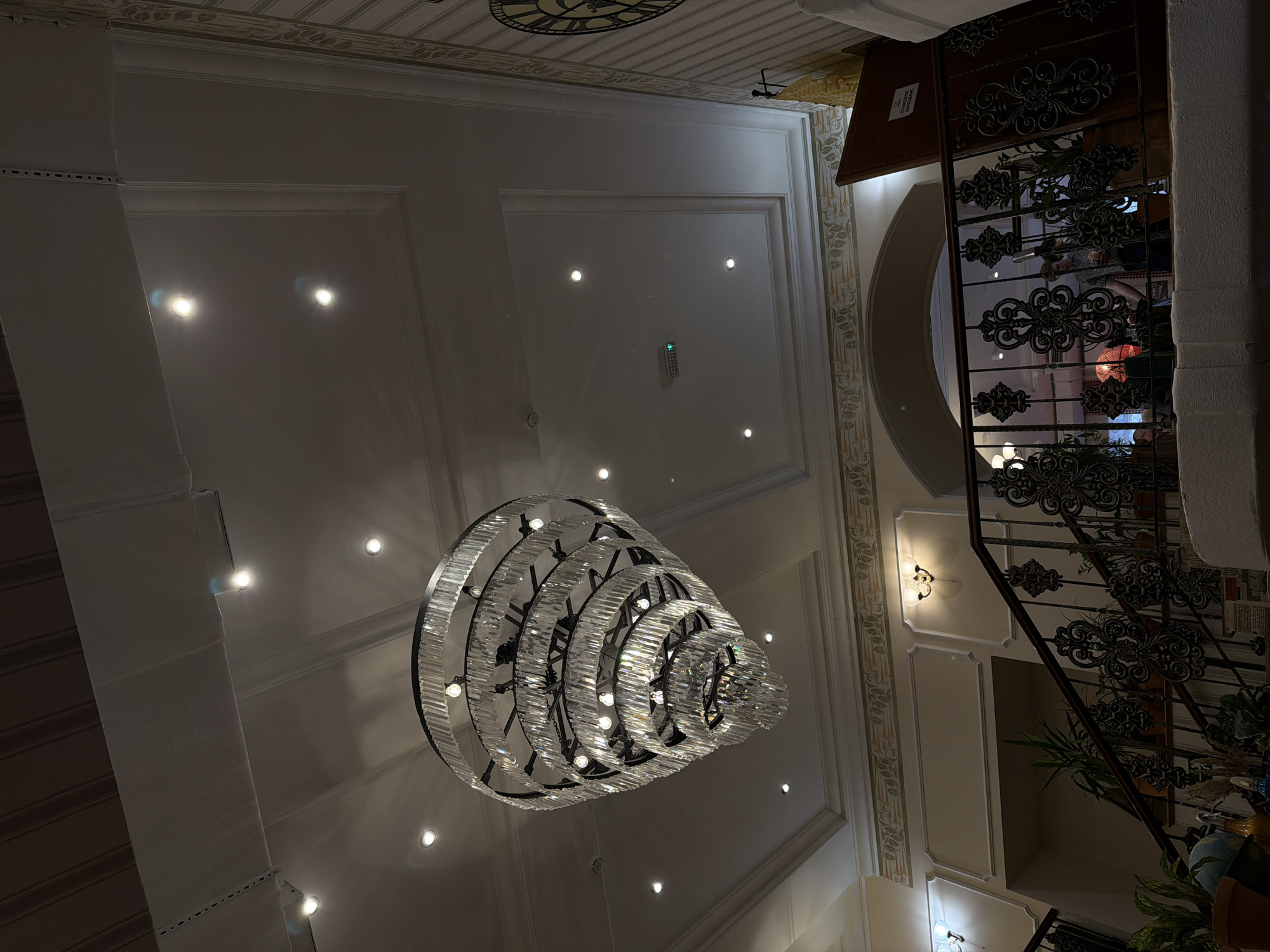 The image captures a grand interior space, likely a hotel lobby or a luxurious building's entrance hall, illuminated primarily by artificial light. A dominant feature is a large, multi-tiered chandelier with crystal-like elements, hanging from a high, ornate ceiling. The ceiling is decorated with recessed spotlights and elaborate moldings, some featuring a subtle leafy pattern. A remote control or thermostat with the partially visible Russian word "ТЕМПЕРАТУРА" (TEMPERATURE) is mounted on the ceiling.
To the right, a grand staircase ascends, featuring intricate black wrought iron railings with decorative scrollwork. Beyond the railings, reflections of indistinct figures can be seen in glass panels, with one showing a blurry person wearing a checkered pattern. Potted plants are placed along the staircase and below it. Several wall sconces provide additional illumination. In the background, a large, traditional clock face is partially visible on an upper wall or ceiling.
The overall impression is one of elegance and grandeur, suggesting an evening or nighttime setting due to the reliance on artificial lighting. The location is specified as Moscow, Russia, though no explicit visual cues within the image directly confirm this. No specific activity is taking place; the image showcases the architectural details and decor of the interior.