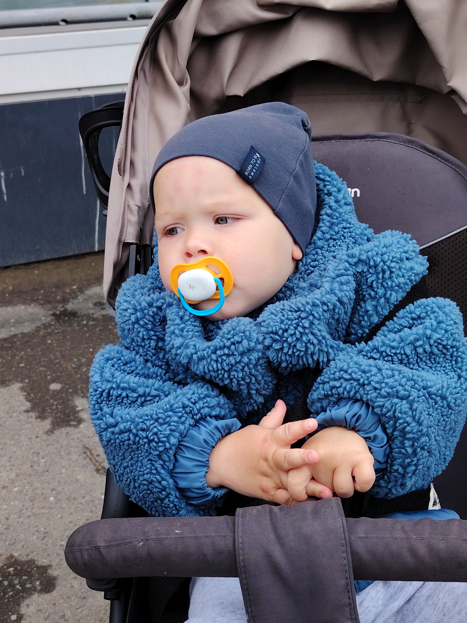 A baby wearing a navy blue beanie and a fluffy blue jacket sits in a stroller with a gray interior and tan canopy. The baby has a pacifier in its mouth, with an orange ring and a blue handle. The baby's hands are folded together in its lap. The background includes a gray paved surface and a building with dark gray and white sections. The location is the city of Chaikovsky, Russia.