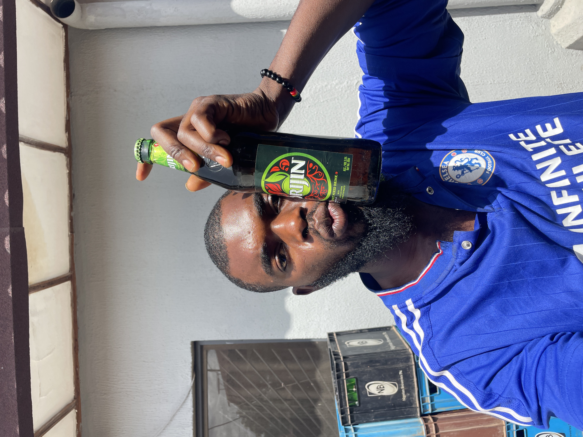 This media file features a close-up, angled shot of a man holding a bottle of "RIJIN" spirit mixed drink. The man, with dark skin and a beard, is wearing a blue Chelsea Football Club jersey. The bottle has a green label with red accents, displaying text like "RIJIN," "SPIRIT MIXED DRINK," "CAN HERB," "FRUIT EXTRACTS," "ALC./VOL 5.5%," and "Net Vol: 60cl." The man is also wearing a black beaded bracelet with red and yellow accents on his wrist.
In the background, stacks of black plastic crates are visible, with one crate showing the "NB" logo and text identifying it as "Nigerian Breweries Plc." A wall with a textured finish and a window with a wire grate are also present. The lighting suggests it is daytime, possibly outdoors or in a well-lit indoor space. The scene appears to be set in Jos, Nigeria, given the location context provided. The man's expression is neutral, and the overall impression is casual.