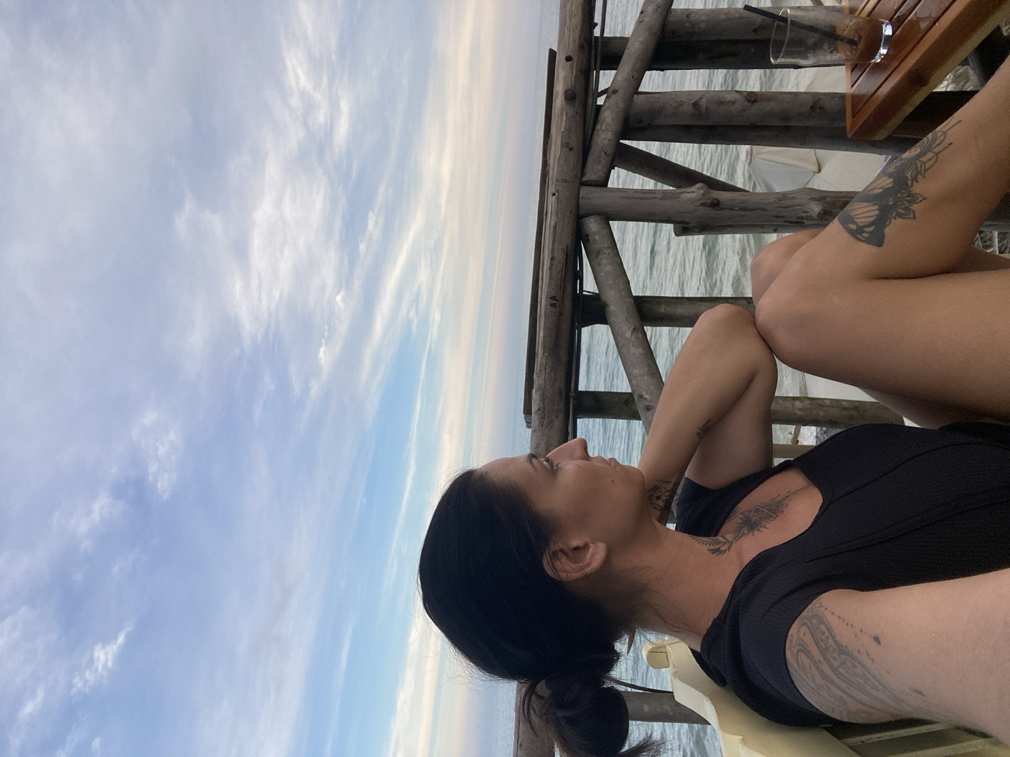 A woman with dark hair tied in a bun, facing right, sits on a light-colored bench, her left arm resting on her lap. She is wearing a black top, and her right arm is bent at the elbow, with her hand behind her head. Tattoos are visible on her left arm, chest, and right leg. In front of her, a wooden table holds a glass with a dark beverage and a straw. Behind her, a wooden railing overlooks the ocean, with gentle waves under a sky filled with wispy clouds. The scene appears to be in Phu Thuy Ward, Vietnam.