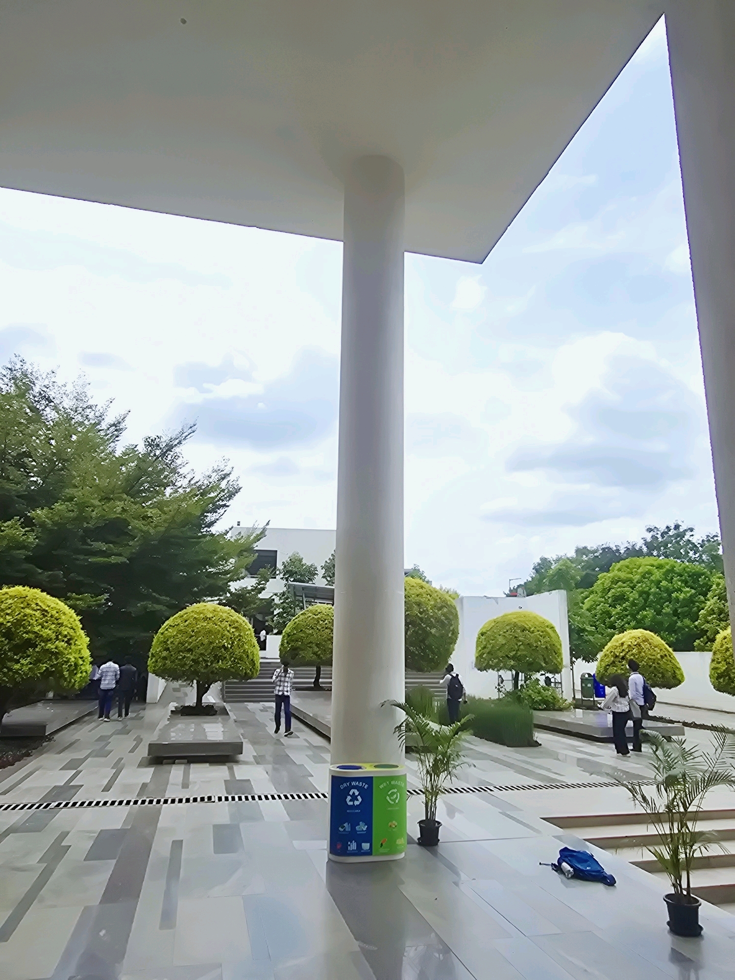 The image shows an outdoor courtyard with modern architecture and landscaping in Ittagalapura, India. The foreground features a light-colored tiled walkway with a decorative pattern of rectangular gray and white tiles. Several potted palm plants are placed on the walkway. To the right, a two-compartment recycling bin is visible, labeled "DRY WASTE" on the left and featuring icons for recycling on the right. A blue backpack lies on the ground near the plants. In the mid-ground, there are several rounded, manicured green bushes and trees. People are walking in the courtyard; two individuals are walking away from the camera on the left, and one person is walking towards the camera on the right. Further in the background, there is a building with white walls and windows, and more trees. The sky above is partly cloudy. The scene appears to be a well-maintained public space.