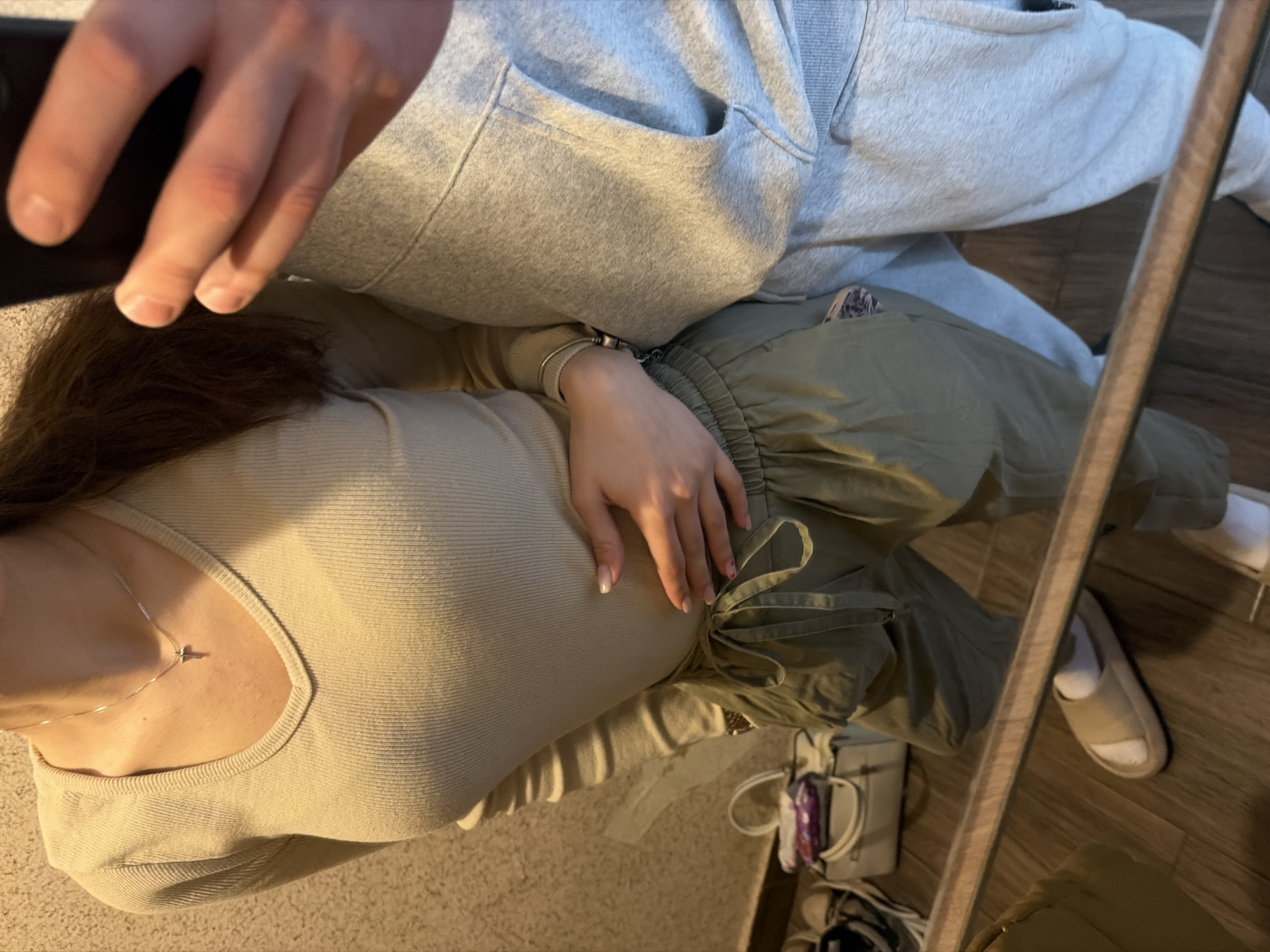 A selfie depicts two individuals in a domestic setting, reflected in a mirror. One person is wearing a light grey cardigan, while the other is wearing a beige top and olive green pants. The setting appears to be indoors, with a floor visible in the reflection. Specific location information in Sofia, Bulgaria is not discernable from the image.