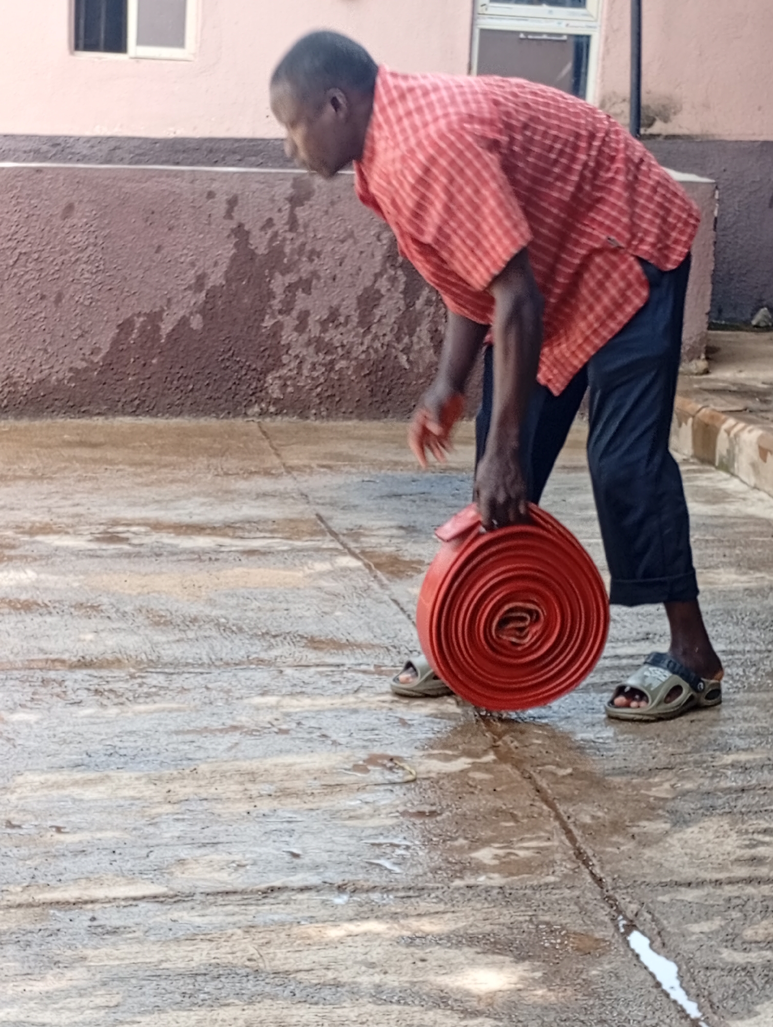 A man, with dark skin and short hair, is shown from the side, bending over to pick up a coiled red hose. He is wearing a red and white checkered shirt and dark blue trousers, with sandals on his feet. The ground is wet and appears to be paved with concrete. In the background, a building with a pinkish-brown wall and a window is visible. The scene suggests a domestic or possibly a work-related setting in Rafin Par, Nigeria. The lighting indicates it could be daytime, and the wet ground may suggest recent rain or cleaning. There is no visible text in the image.
