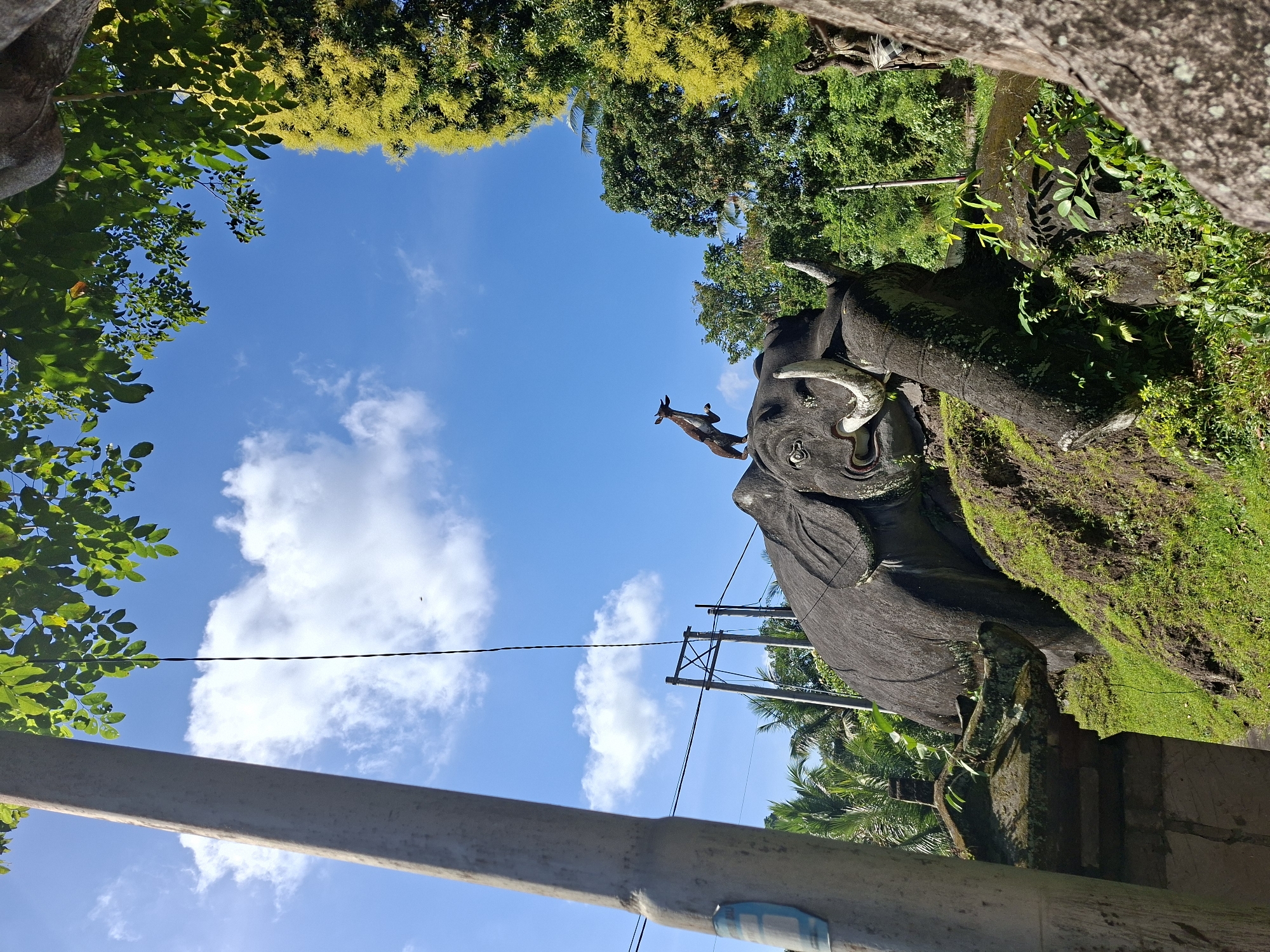 This image depicts a stone sculpture of an elephant, possibly a temple guardian or a monument, situated outdoors in Banjarangkan, Indonesia. The elephant sculpture is large, dark gray, and appears to be resting on a moss-covered hillock. It has prominent tusks and an open mouth. Perched on the elephant's head is a smaller, dark sculpture of an animal, possibly a deer or a goat, in a leaping pose. The background is dominated by a bright blue sky with scattered white clouds. Lush green foliage and trees surround the elephant, with some palm trees visible in the distance. A utility pole with electrical wires stands to the left of the elephant. The lighting suggests it is daytime, with sunlight casting shadows.