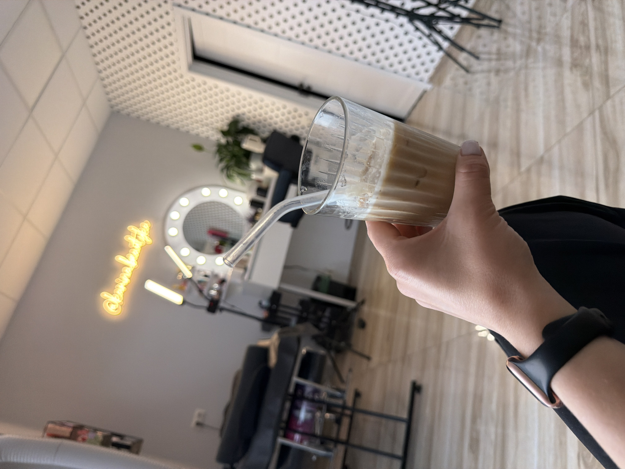 A person's hand holds a glass of iced coffee with a straw. The background appears to be a salon or beauty studio, with a lighted vanity mirror, a treatment couch, and a neon sign on the wall that reads "@kamirabeauty." The overall setting suggests a commercial establishment. The image does not contain any information that would allow it to be located in Riga, Latvia.