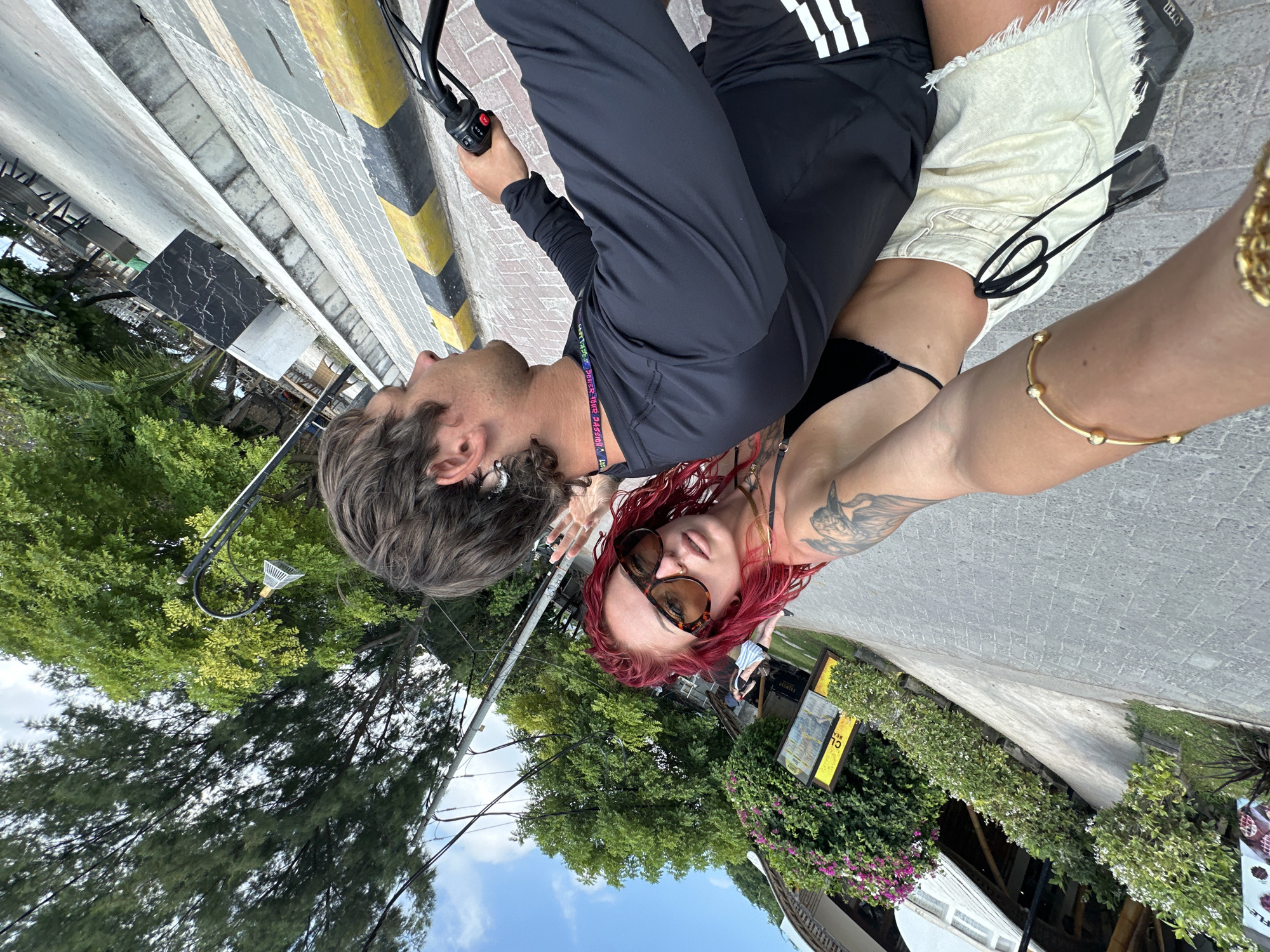 A couple is riding an electric bike on a street in what appears to be a tropical location. The man is driving, wearing a black Adidas tracksuit and a lanyard. The woman beside him has red hair, sunglasses, a black bikini top, a tattoo on her arm, and a gold bracelet. They are surrounded by lush greenery and a partially visible building on the right side of the frame. A lamp post and utility wires are visible among the trees. The street is paved with concrete or stone blocks, and the curb has alternating black and yellow stripes. A black marble table is situated near the curb. In the background, a blue sky is visible between the trees. A sign is partially visible on the right. The city location is unknown but is in Indonesia.