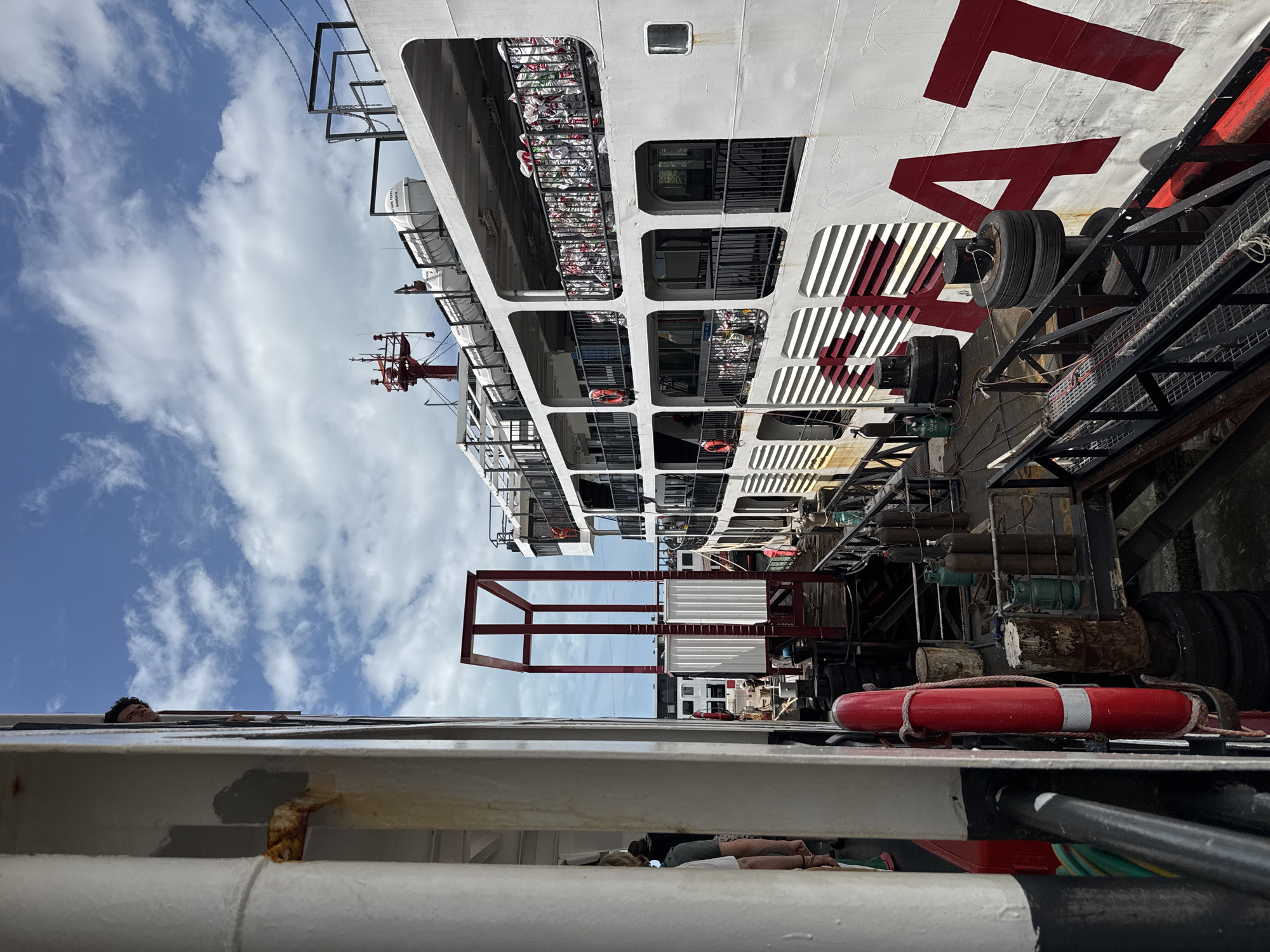 The image captures a docking scene at a ferry terminal. A large white ferry with red lettering, possibly a brand name, is moored alongside a smaller vessel or pier structure. The ferry has multiple decks visible, with railings and windows. Life preservers and what appear to be stacked bags or cargo are visible on one of the ferry's decks.
In the foreground, a portion of a white vertical structure, likely part of another vessel or dock, occupies the left side of the frame. A red life preserver is attached to this structure. A person is partially visible in the upper left quadrant, looking out from an opening in the structure.
The background features a blue sky with scattered white clouds. A red metal framework structure stands between the two vessels. The ground area appears to be a walkway or pier, with various objects like tires, canisters, and wooden posts scattered around. The overall impression is one of a busy maritime transportation hub.