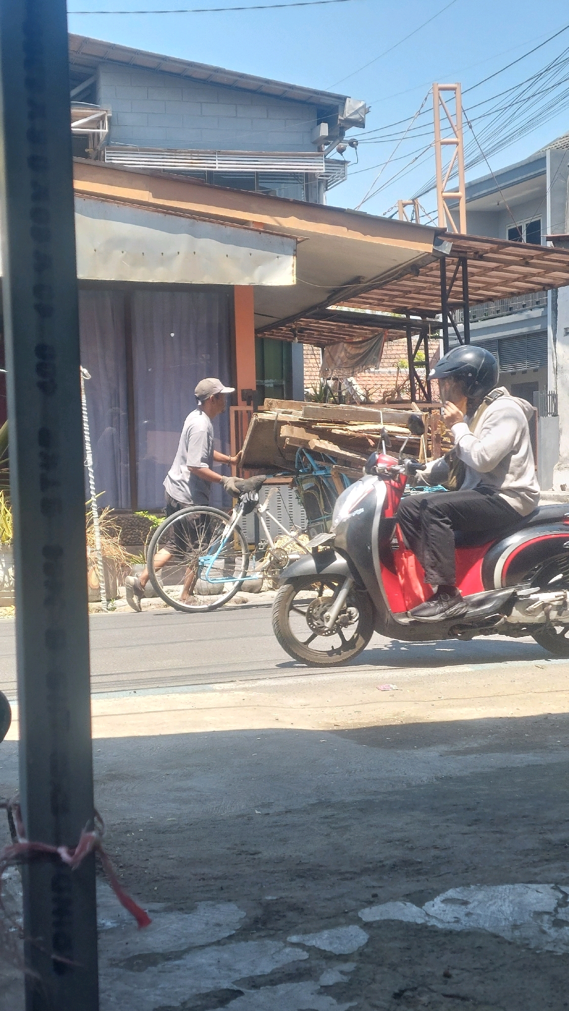 In Sidoarjo, Indonesia, under a bright, sunny sky, a man wearing a grey t-shirt, dark shorts, and a cap pushes a bicycle loaded with wooden planks. The bicycle has a blue frame and a front basket. To the right, a person wearing a grey hoodie, dark pants, and a black helmet rides a red and black scooter, holding what appears to be a mobile phone to their ear. The scene unfolds on a paved road in front of buildings with awnings and overhead electrical wires. The foreground features a blurred vertical structure.