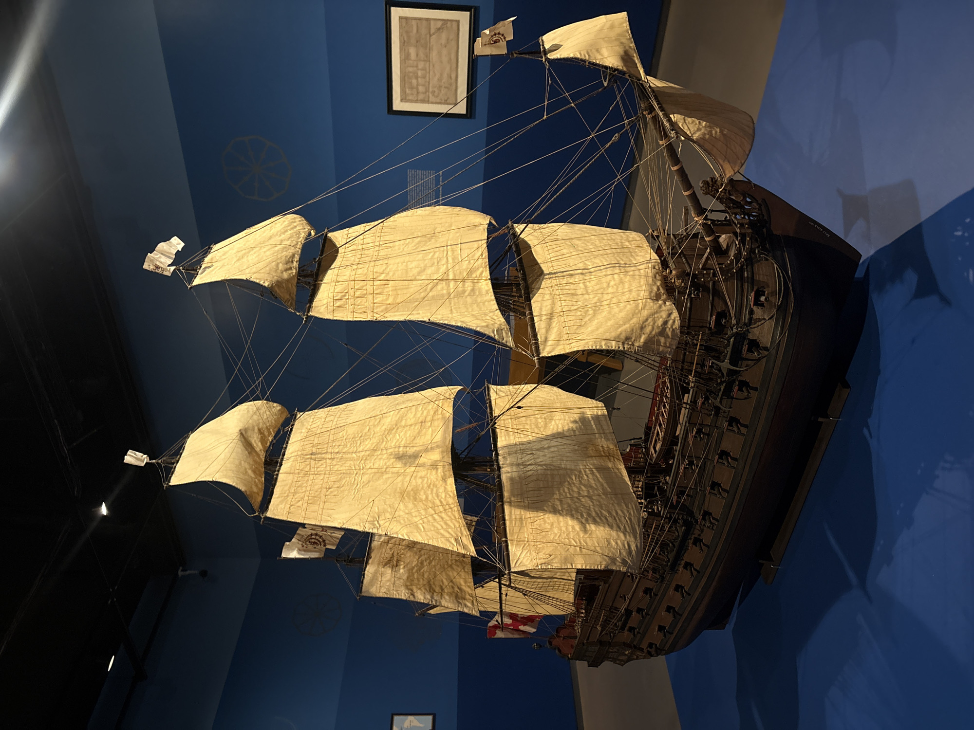 A model ship is on display at the Museo Naval Mexico in Mexico City, Mexico. The model is of a sailing ship with multiple masts and sails. The sails are cream colored and appear to be made of fabric. There is rigging connecting the masts and sails. The hull of the ship is dark brown or black and has multiple decks. The ship is displayed against a blue wall. There is a framed print hanging on the wall behind the ship. There are faint circular designs on the blue wall. The lighting is bright and the image is taken from a slightly low angle.