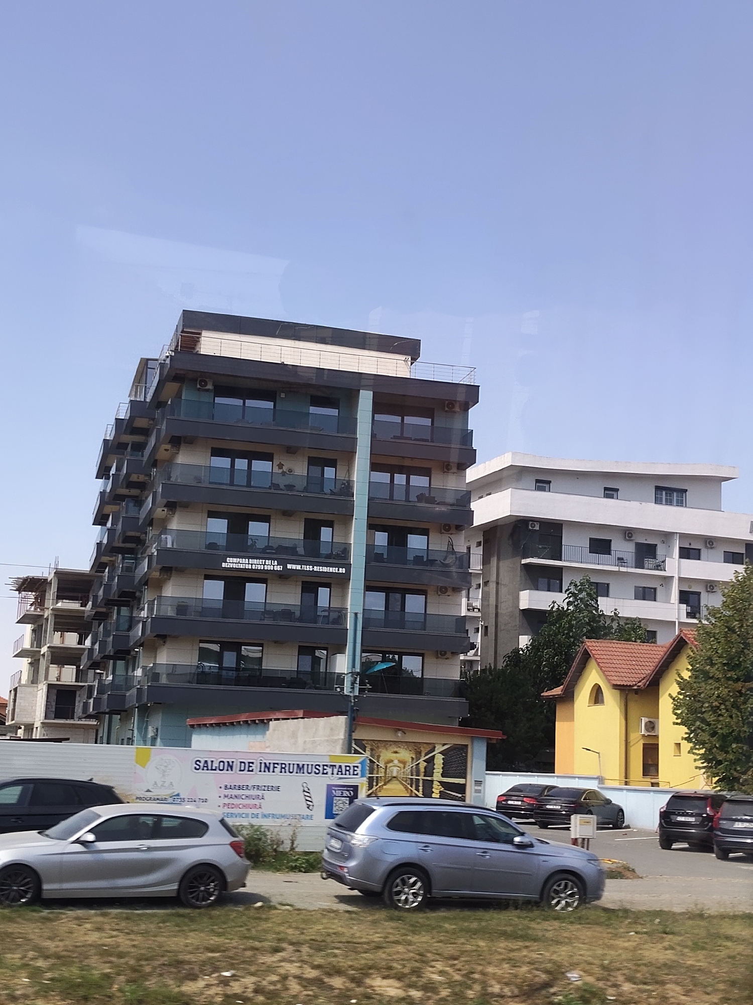 The image depicts a street scene in Corbu, Romania, featuring modern apartment buildings under a clear blue sky. In the foreground, several cars are parked on a paved area adjacent to a grassy strip. A silver hatchback is prominent on the left, while a light blue SUV is positioned in the center. Further back, darker sedans and SUVs are visible.

A large building with a dark facade and balconies is the dominant structure on the left side of the image. It has multiple stories with glass-fronted balconies. A banner advertisement is visible on one of the lower floors of this building, reading "CUMPARA DIRECT DE LA DEZVOLTATOR 0755 050 657 WWW.TLSS-RESIDENCE.RO". To the right of this building, a white apartment complex with multiple windows and balconies is present. A smaller, yellow building with a red-tiled roof is situated in the lower right quadrant, and a building under construction is visible to the left of the main apartment buildings. A sign for "SALON DE INFRUMUSETARE" (Beauty Salon) is displayed on a banner across the lower part of the image, listing services such as "BARBER/FRIZERIE", "MANICHIURA", "PEDICHIURA", and "SERVICII DE INFRUMUSETARE". The environment appears to be a residential or mixed-use urban area. There are no people clearly visible in the scene.