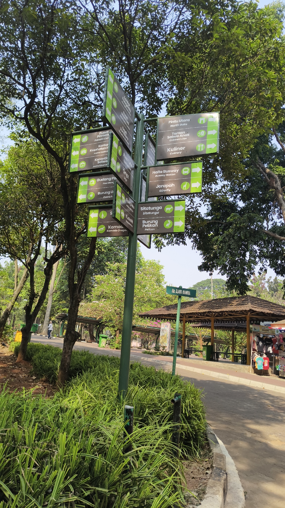 A directional signpost stands in a park-like setting with lush green foliage. The signpost is a dark green metal pole with multiple rectangular signs attached at various angles. Each sign has white text and icons indicating destinations and directions, with arrows pointing left or right. Some of the legible signs include "Pintu Utara" (North Gate), "Taman Satwa Anak" (Children's Zoo), "Kuliner" (Culinary), "Halte Busway" (Busway Station), "Jerupah" (Giraffe), "Situtunga" (Sitatunga), and "Burung Pelikan" (Pelican). The background features tall trees with dense green leaves, suggesting a sunny day. In the distance, there are shaded structures with wooden roofs, pathways, and some people walking. The scene appears to be a recreational area, likely a zoo or a large public park, possibly located in South Jakarta, Indonesia, given the Indonesian text on the signs.
