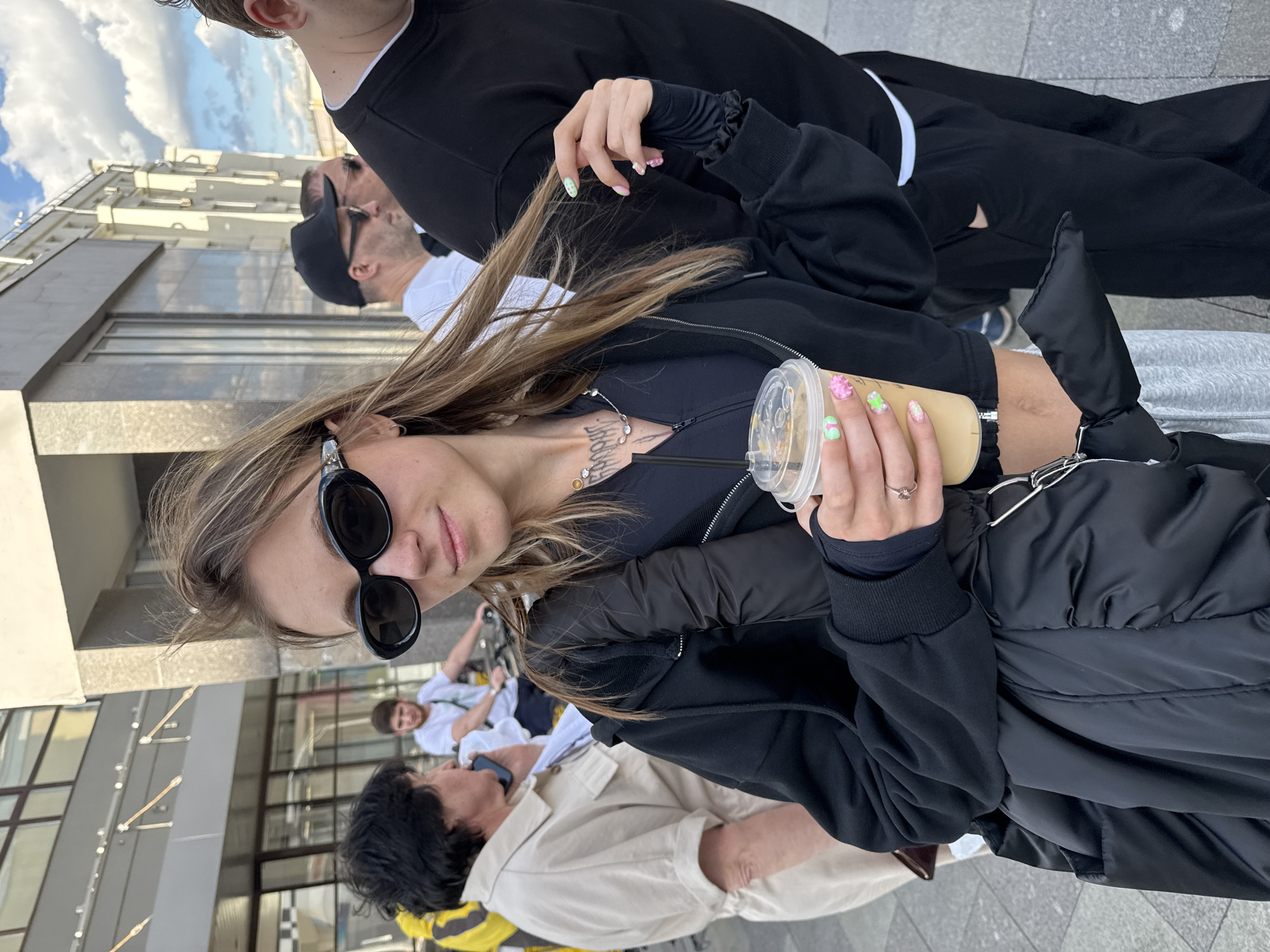 The image shows a group of people, seemingly outdoors on a sidewalk in Moscow, Russia. In the foreground, a young woman with blonde hair pulled back and wearing sunglasses, a black zip-up jacket over a black top, and gray sweatpants is holding a clear plastic cup filled with a light-colored beverage. Her nails are painted with colorful designs.
Behind her, a person wearing a black t-shirt and black pants is partially visible. Further back, a man wearing a black baseball cap and sunglasses is seen with a man in a white t-shirt. Another person in a light-colored jacket is also present in the background. The group appears to be walking or standing together, possibly during a casual outing.
The background includes a modern building with large windows, suggesting an urban environment. The sky is partly cloudy, indicating it might be daytime, though the exact time is not discernible. The overall atmosphere seems relaxed and informal. There is no visible text in the image.