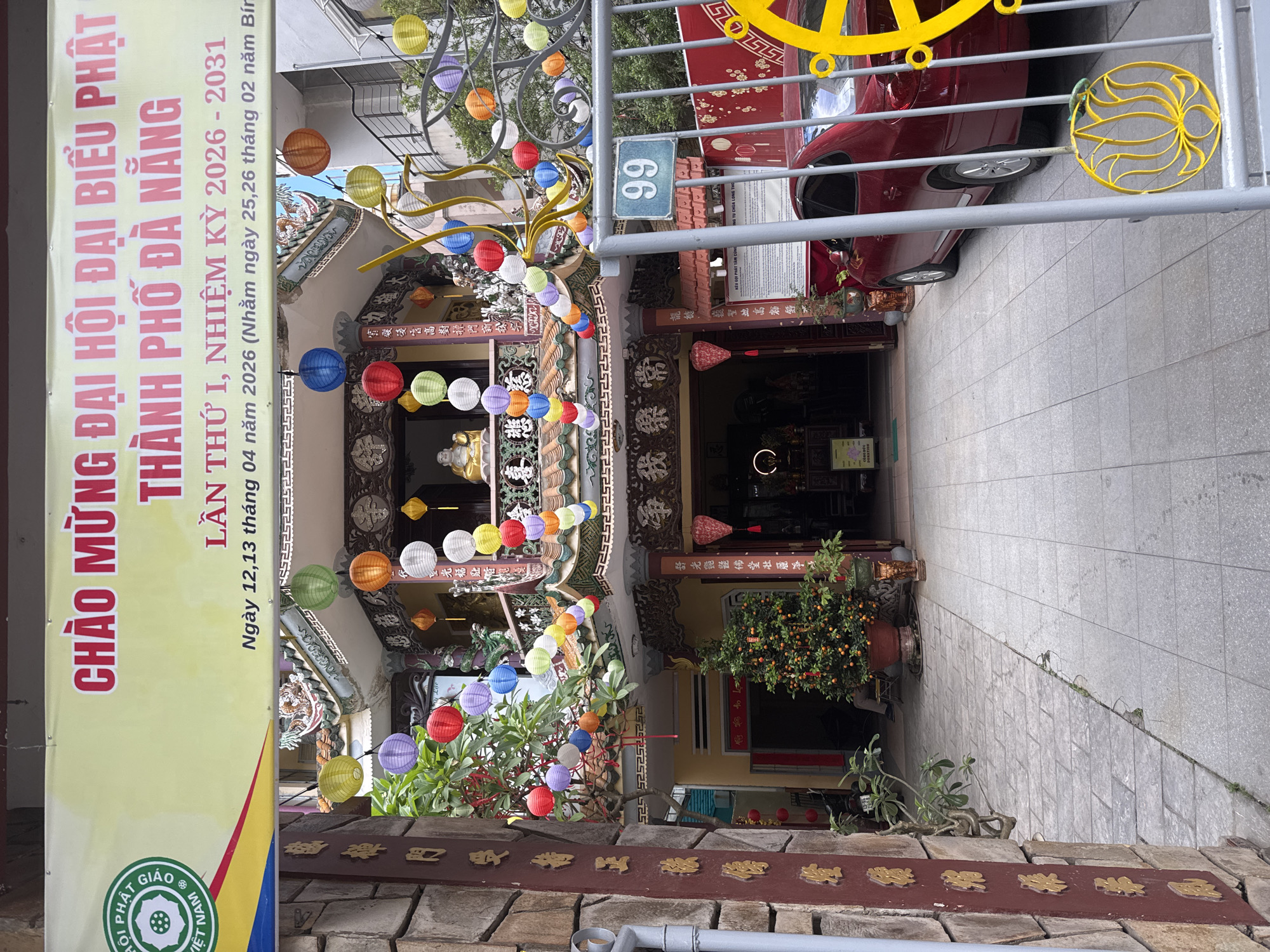 The image shows the entrance to a building, possibly a Buddhist temple, decorated with colorful lanterns. A banner to the left of the entrance reads "Chao Mung Dai Hoi Dai Bieu Phat Thanh Pho Da Nang", indicating it's a celebration for the Buddhist community of Da Nang. The banner also mentions the period of 2026-2031, which is likely related to the event being celebrated. The building number "66" is visible on a sign above the entrance. There's a red car parked in front of the building. The overall scene appears to be taken on a street in Da Nang, Vietnam.