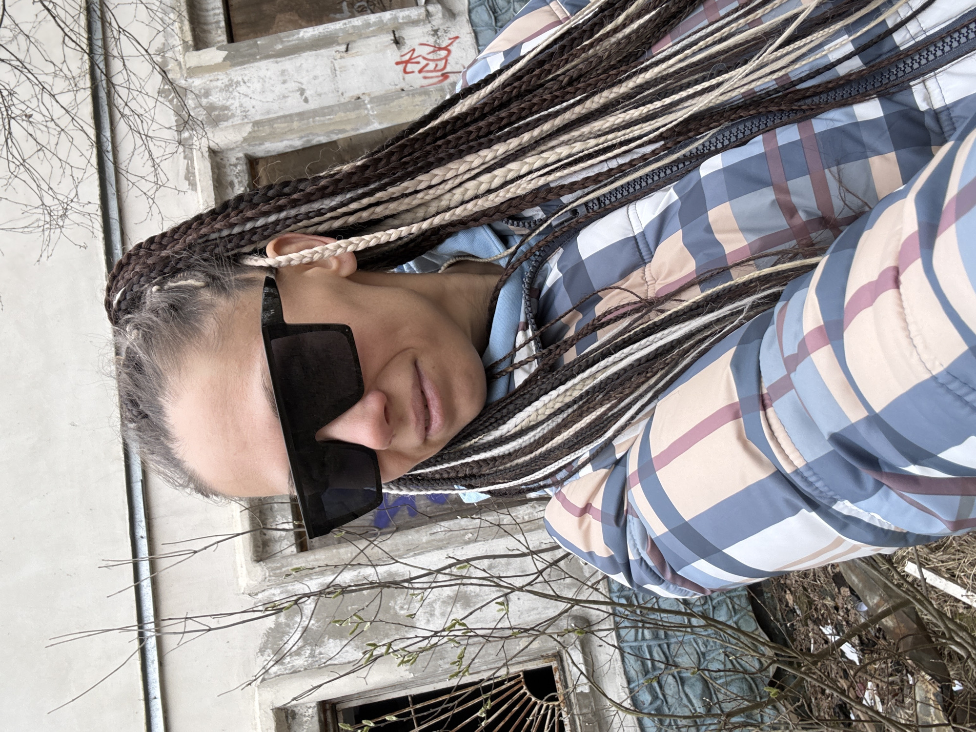 An individual is captured in a rotated selfie, focusing on their head and upper torso against a backdrop of a derelict building and tree branches. The person sports long, multi-toned dreadlocks in shades of dark brown, beige, and white/grey. They wear large, dark, rectangular sunglasses that obscure their eyes. Their attire includes a puffer jacket featuring a plaid pattern with hues of blue, light blue, beige, and a muted reddish-purple, fastened with a dark zipper. A lighter blue garment is visible underneath.
The background reveals an old, weathered concrete or stucco building, showing clear signs of neglect and decay, including empty or broken window frames with some visible twisted metal. Numerous bare tree branches, some with tiny green buds indicative of early spring, are prominent throughout the scene. Faint red Cyrillic graffiti is discernable on one of the building's walls.
The lighting suggests daytime, possibly under an overcast sky, given the soft, even illumination. The overall scene conveys an urban or semi-urban environment in disrepair. The person's visible expression appears neutral. This setting aligns with the provided location context of Rusanovo, Russia.