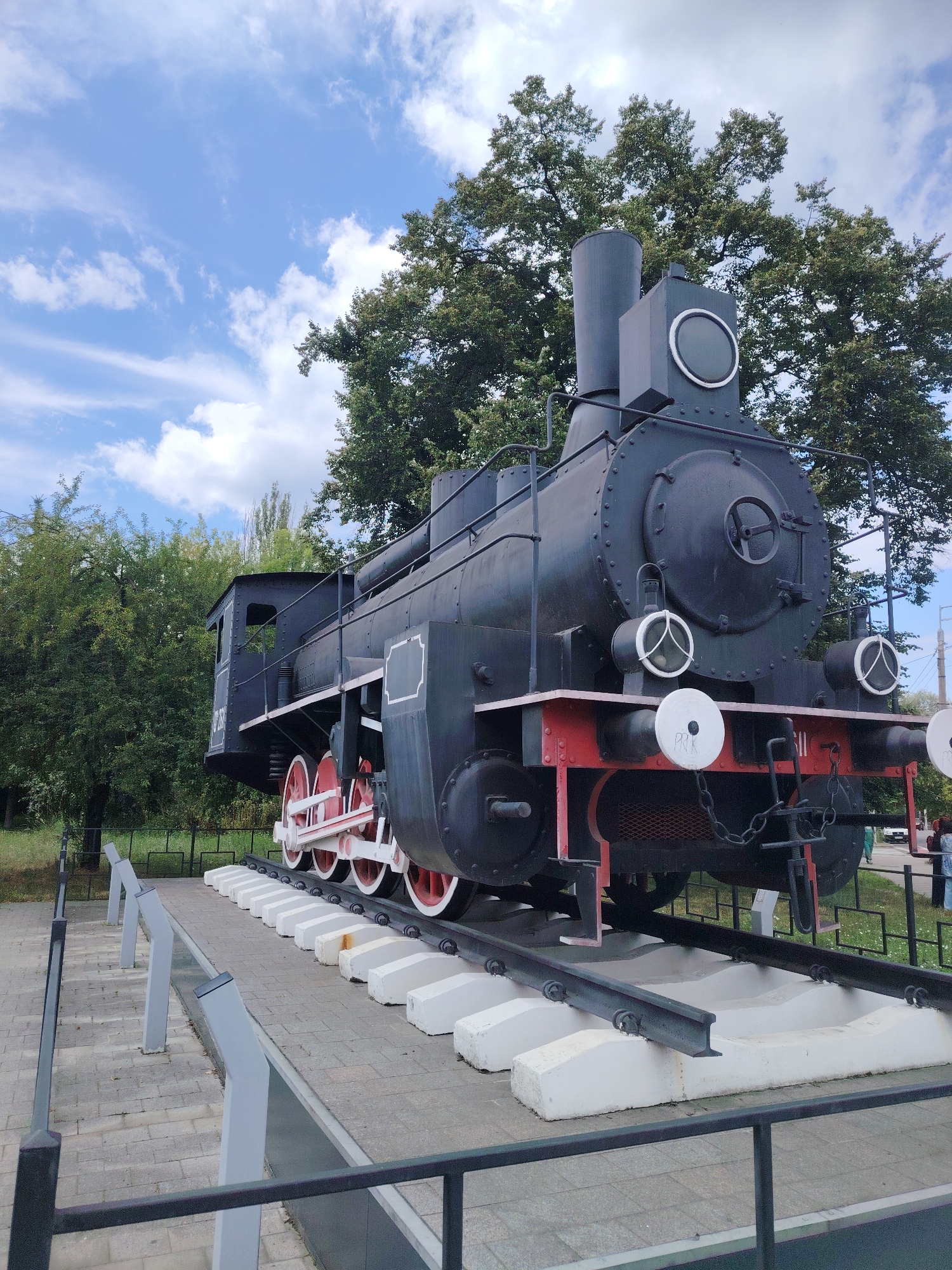 An old black steam locomotive, likely a monument, is displayed outdoors on a section of track. The locomotive is positioned at an angle, with its front facing the viewer. It features large red-rimmed wheels and a prominent smokestack. The surrounding environment includes lush green trees, a blue sky with scattered clouds, and a paved area with a low black metal fence. There are informational display stands along the paved area in front of the locomotive. In the background, a few people are visible near a road, and there is a white car. The scene appears to be in a public space, possibly a park or memorial site. While the specific location in Kyiv, Ukraine is not visually identifiable from the image alone, this type of display is common in historical sites or parks within the city.