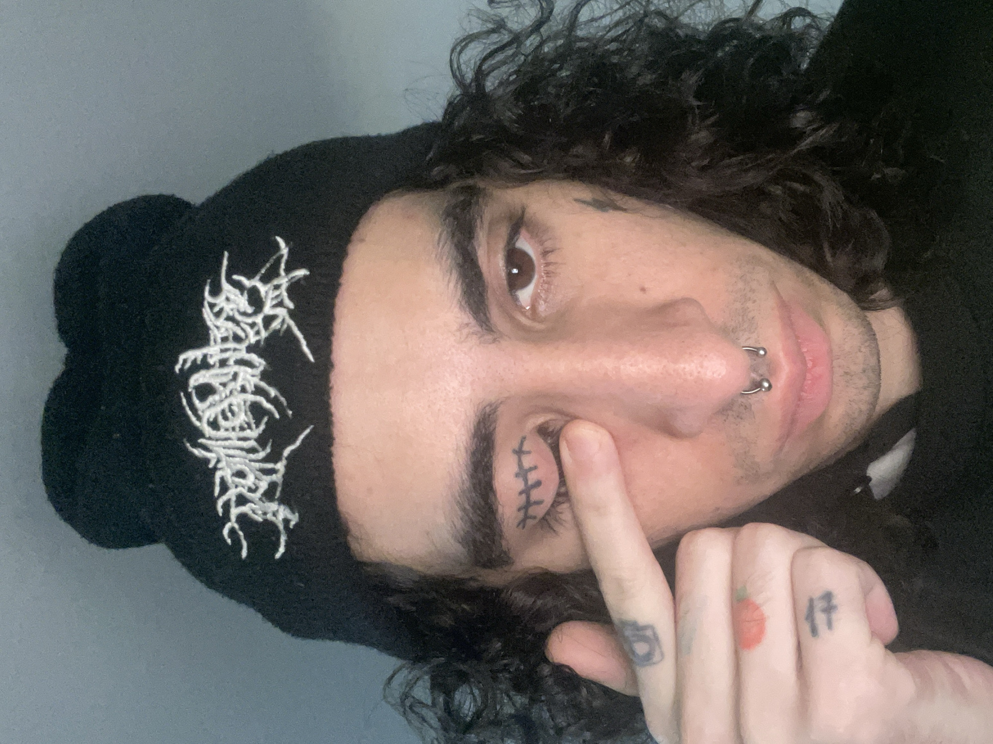 A close-up, angled shot depicts a person with dark, curly hair wearing a black beanie with a light-colored, illegible design. The person has a nose ring and tattoos on their fingers, including numbers and an apparent strawberry. One finger is held near the person's closed left eye, which has a tattoo resembling stitches. The background is a plain light gray. The location is not discernible from the image.
