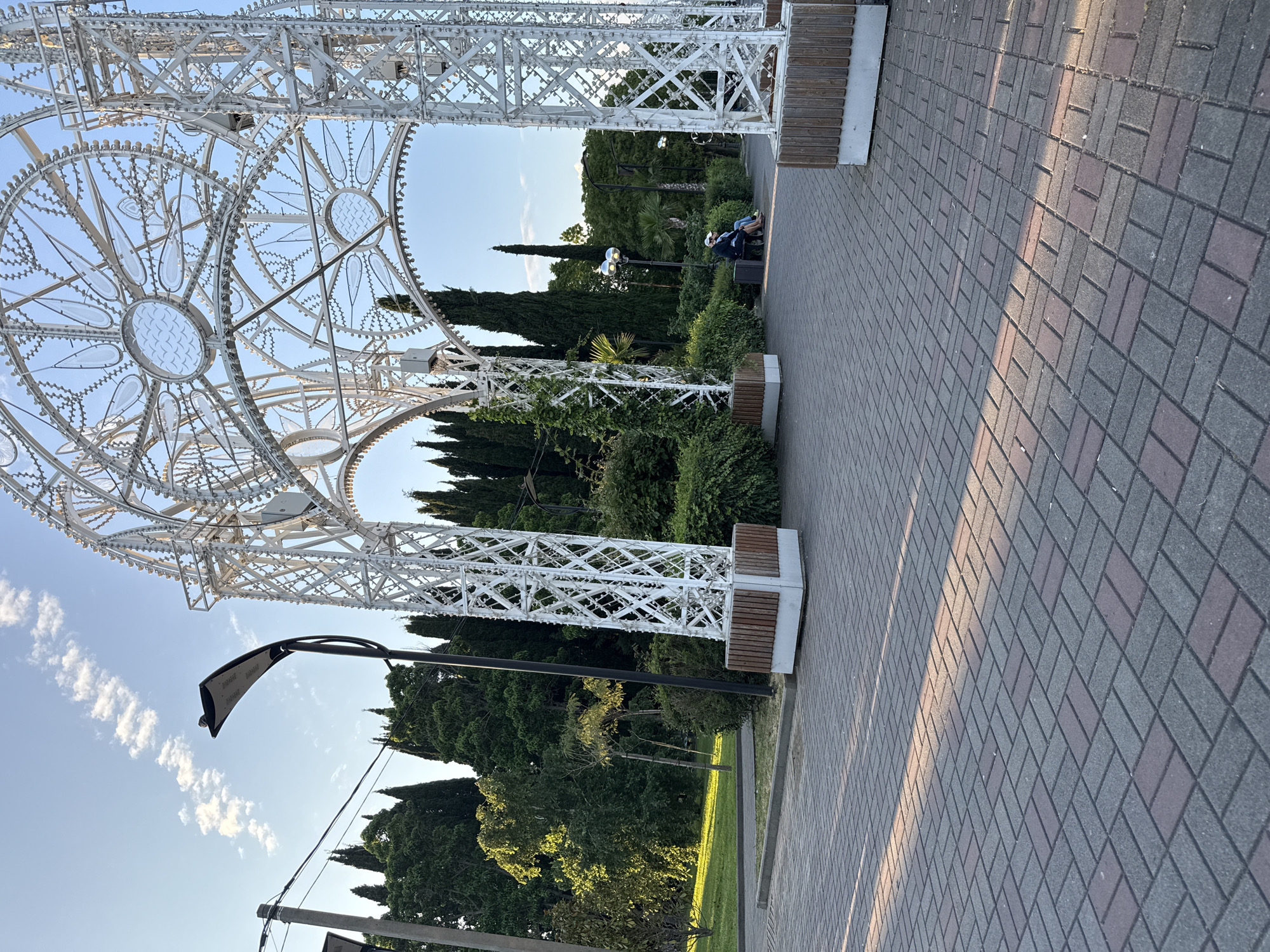 The image depicts an outdoor scene in Sochi, Russia, likely during the late afternoon or early evening due to the angled sunlight casting shadows. The foreground features a brick-paved pathway arranged in a herringbone pattern, with dappled sunlight creating diagonal lines across the surface.
Dominating the right side of the frame is a large, white, ornate metal archway, possibly an art installation or decorative structure. It is constructed with intricate geometric patterns and appears to be illuminated by numerous small lights. The archway has a substantial base with wooden slats.
To the left of the archway, there is a pathway leading into a lush green park area. This area is densely populated with tall, dark green trees, including cypress-like evergreens and broader deciduous trees. In the midground, on a bench under the archway, two people are seated, facing away from the camera and appearing to be in conversation. One individual wears a blue shirt and a white cap, while the other wears a darker top and a white cap.
Further into the park, additional streetlights are visible, some with a modern, curved design. The sky above is a clear blue with scattered, wispy white clouds. The overall impression is one of a well-maintained public space or park within a city, designed for leisure and visual appeal.