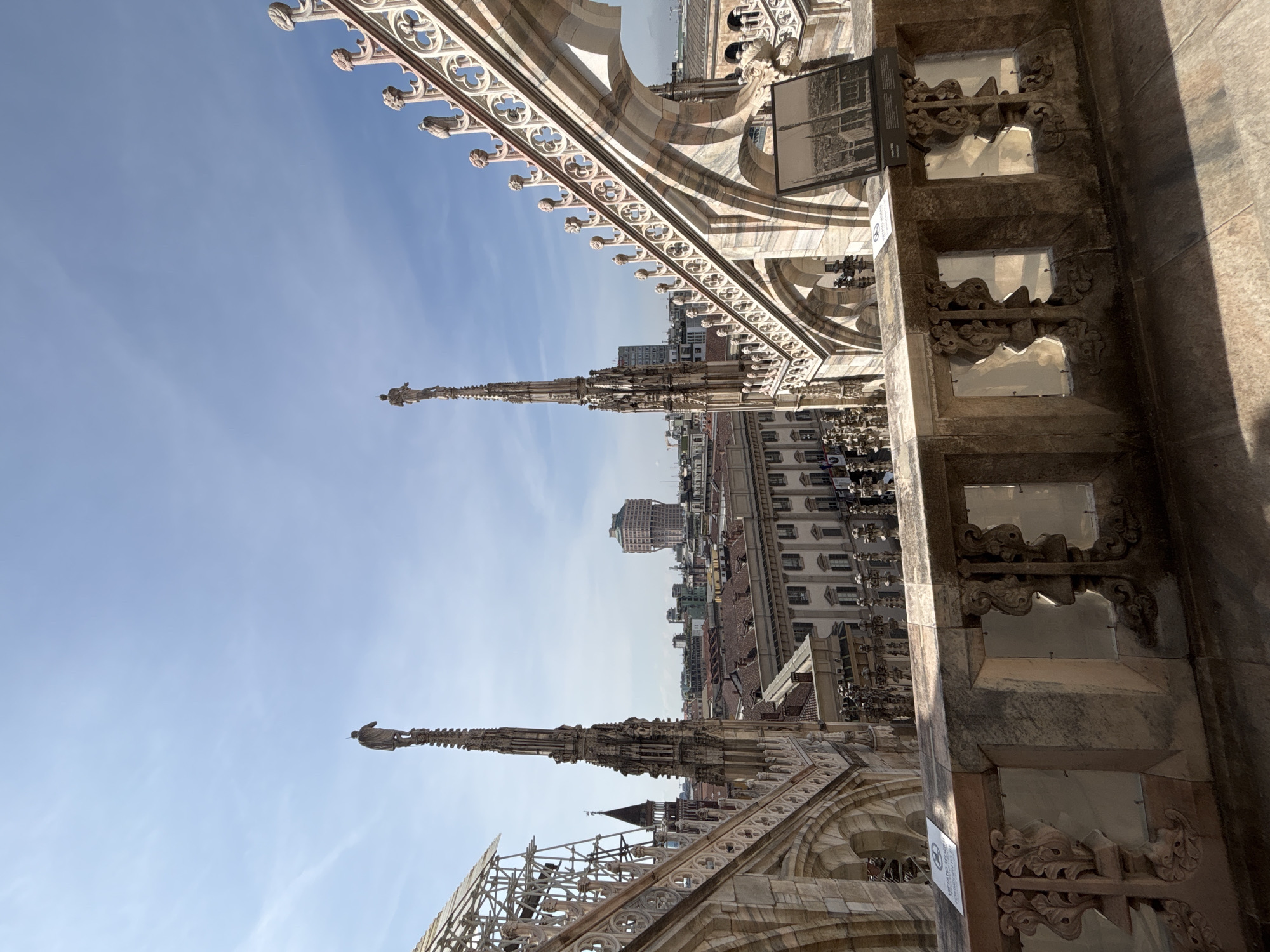 The image shows an upward view from the rooftop of the Milan Cathedral (Duomo di Milano) in Milan, Italy. Elaborate Gothic architectural details of the cathedral are visible, including spires, statues, and ornate stonework. In the background, city buildings are visible against a clear blue sky. The photo is taken in daylight with the cathedral's stonework appearing weathered and aged. There are no people clearly visible in the image.