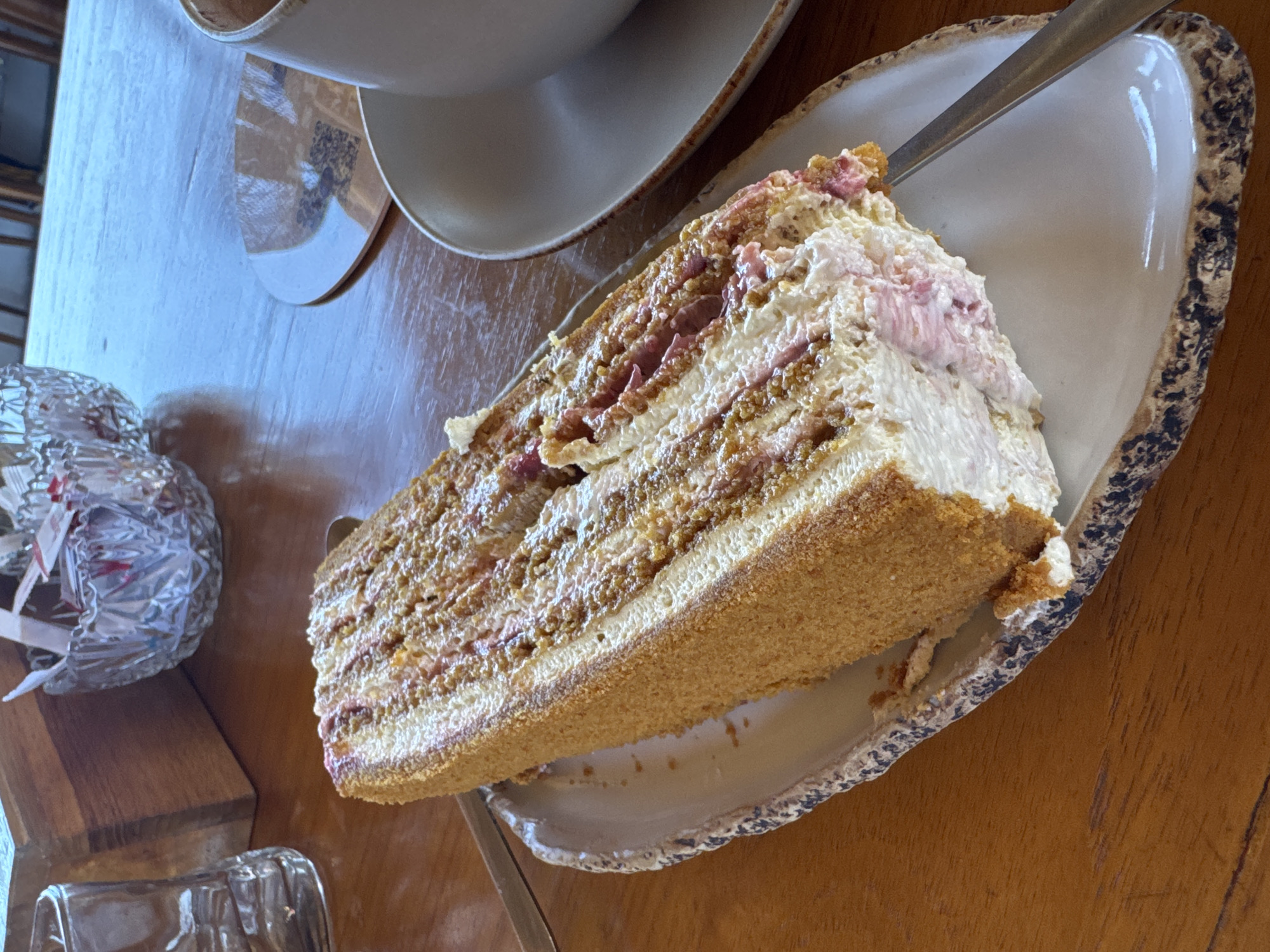 A slice of layered cake rests on a plate with a textured rim, with a fork alongside it. The cake features multiple layers of filling and a crumb-coated exterior. A cup and saucer are visible in the background, along with a small package wrapped in plastic and a drinking glass. The scene is set on a wooden table. The location is Kuta Selatan, Indonesia.