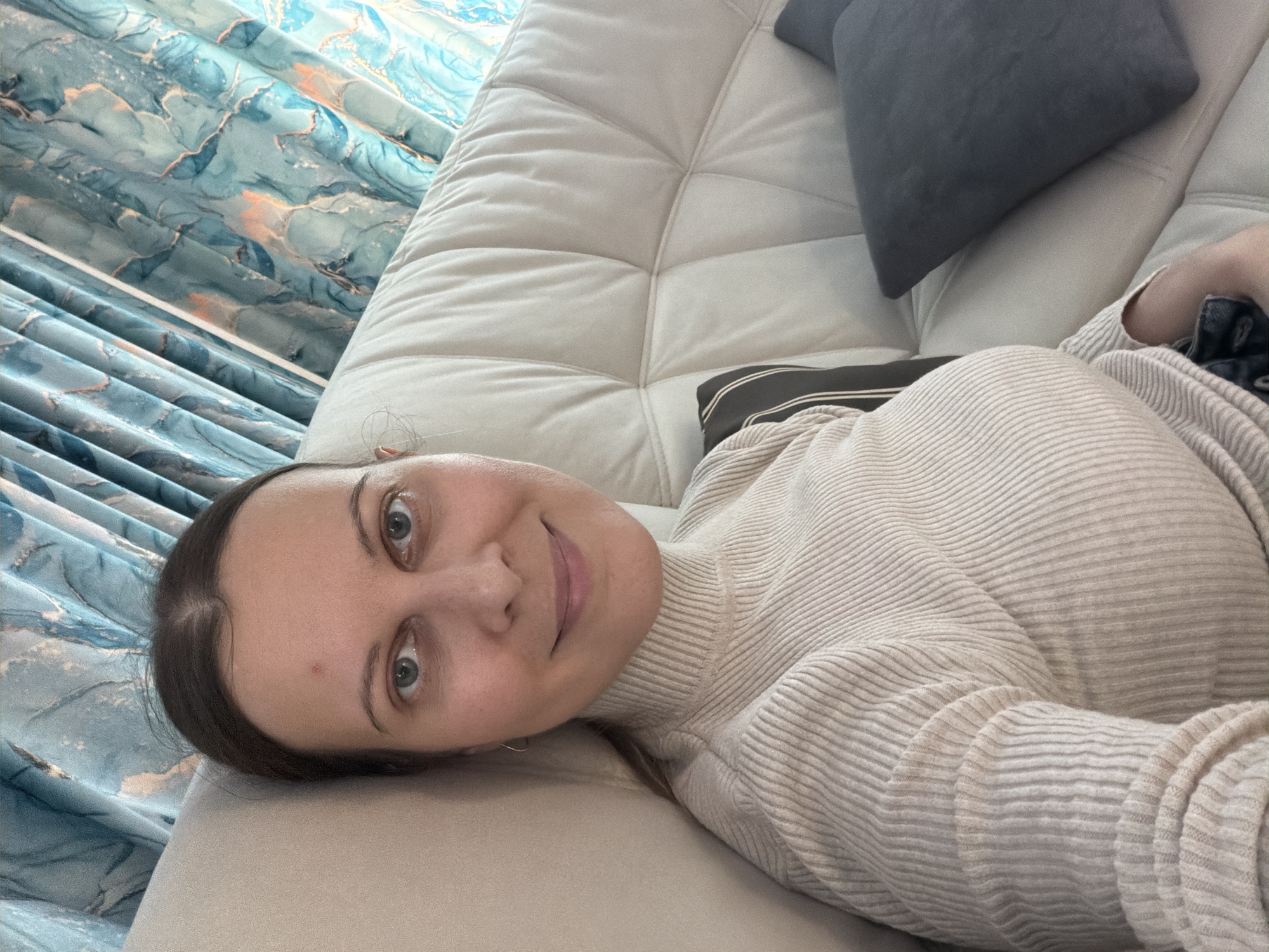 A medium shot captures a woman lying on her side on a light-colored sofa, looking directly into the camera with a calm, slightly smiling expression. She has light-colored eyes and dark hair, with a small reddish mark visible on her forehead. She is wearing a light beige or off-white ribbed turtleneck sweater.
The sofa is upholstered in a tufted light fabric, possibly beige or off-white. In the background, to the upper left, patterned curtains with blue and teal tones and abstract gold or orange marble-like designs are visible, suggesting natural light from a window. A dark gray square pillow rests on the sofa behind her, and a striped pillow is partially visible near her body. A glimpse of dark denim fabric is seen near her right arm.
The image conveys a relaxed indoor setting, likely a living room or lounge area. The woman appears comfortable and at ease. The scene takes place in Старое Михайловское, Russia. There is no visible text in the image.