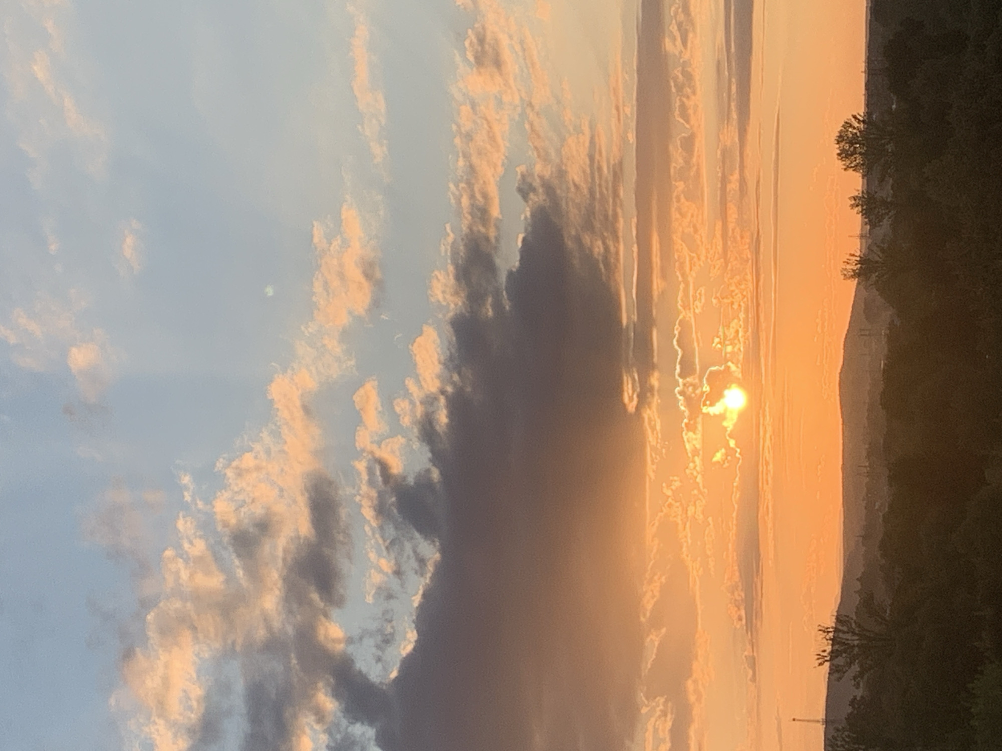 The image shows a sunset over a landscape that appears to be on the outskirts of Kolomyia, Ukraine. The sky is filled with clouds, some of which are illuminated by the setting sun with hues of orange and pink. The sun itself is visible as a bright orb peeking through the clouds on the right side of the frame. Below the sky, a silhouetted treeline forms the horizon, with some hints of distant buildings and structures, possibly industrial, visible in the far distance. The overall scene is peaceful and captures the natural beauty of a sunset.
