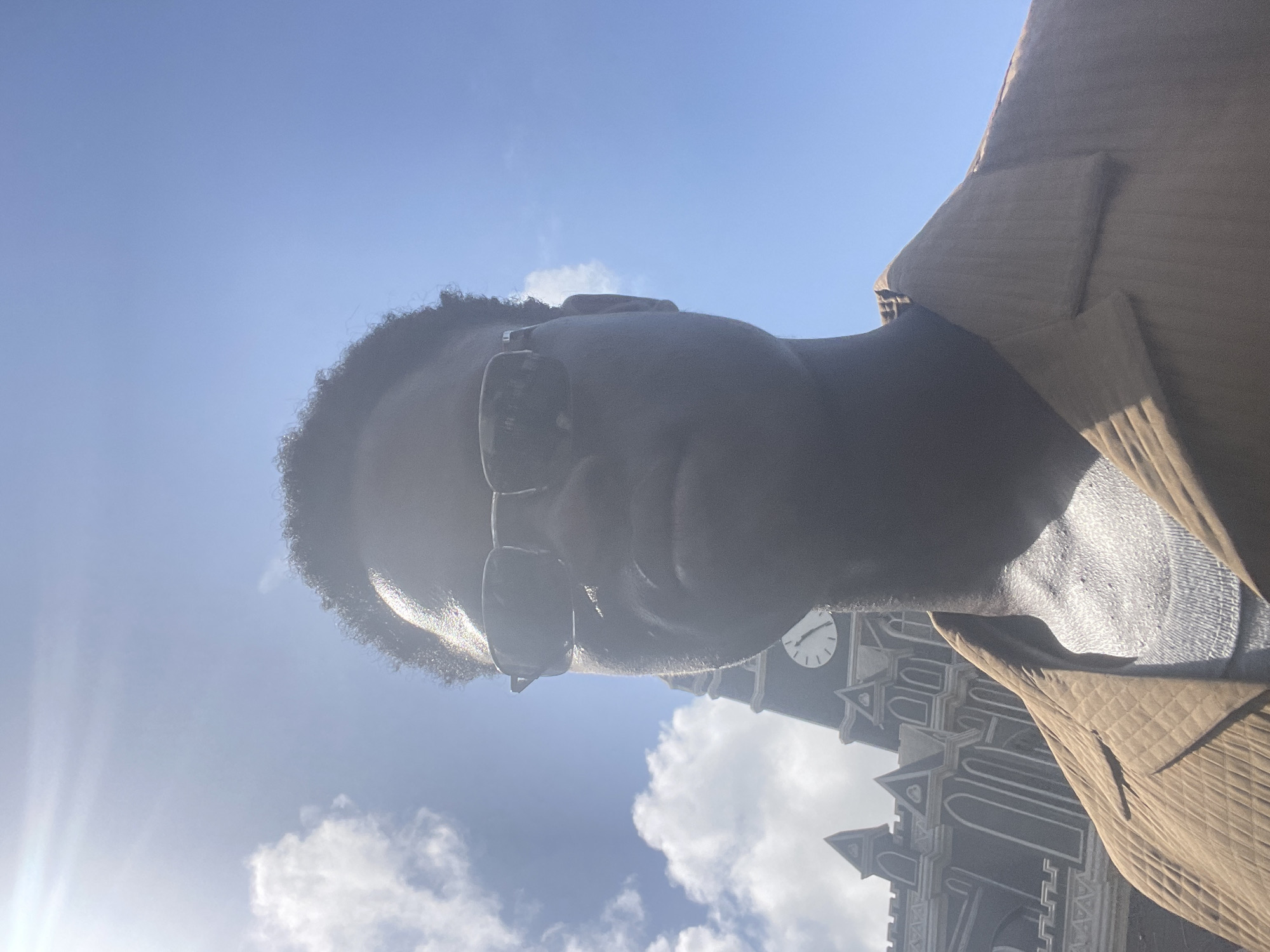 The image is an upward-angle selfie of a person wearing sunglasses and a brown corduroy jacket. In the background, partially obscured by the subject, is a building with architectural details suggestive of a cathedral or church. A clock face is visible on one of the towers. The sky is blue with some clouds. The bright light suggests a sunny day. It is not possible to confirm that the location is Lagos, Nigeria, from the image alone.