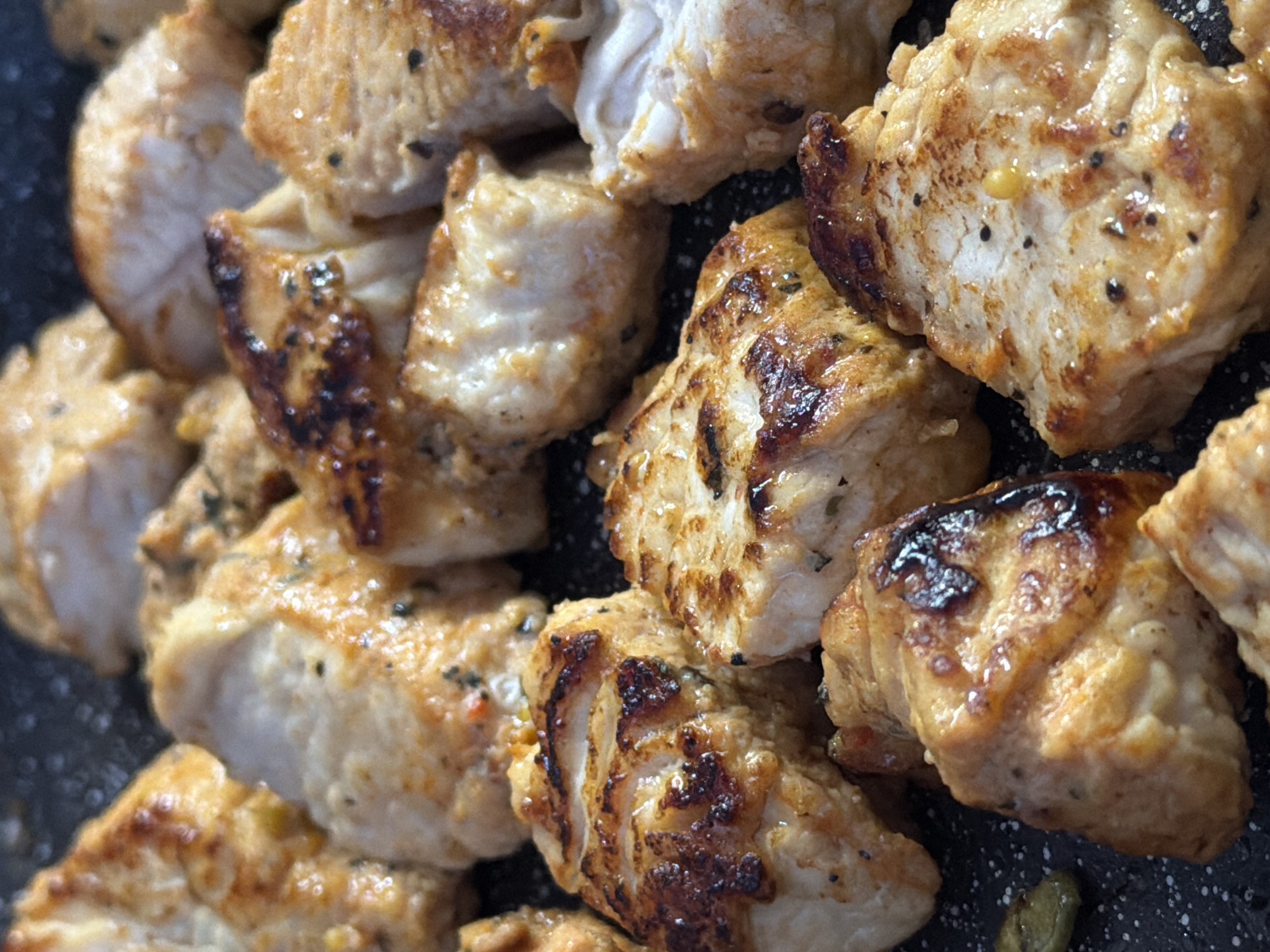 The image shows a close-up of cooked chicken pieces. The pieces are browned and appear to have been pan-fried or grilled. Some pieces show distinct grill marks. The chicken appears to be seasoned with pepper and herbs. The cooking surface is dark with light-colored speckles.
There is no indication of a location in Izhevsk, Russia.