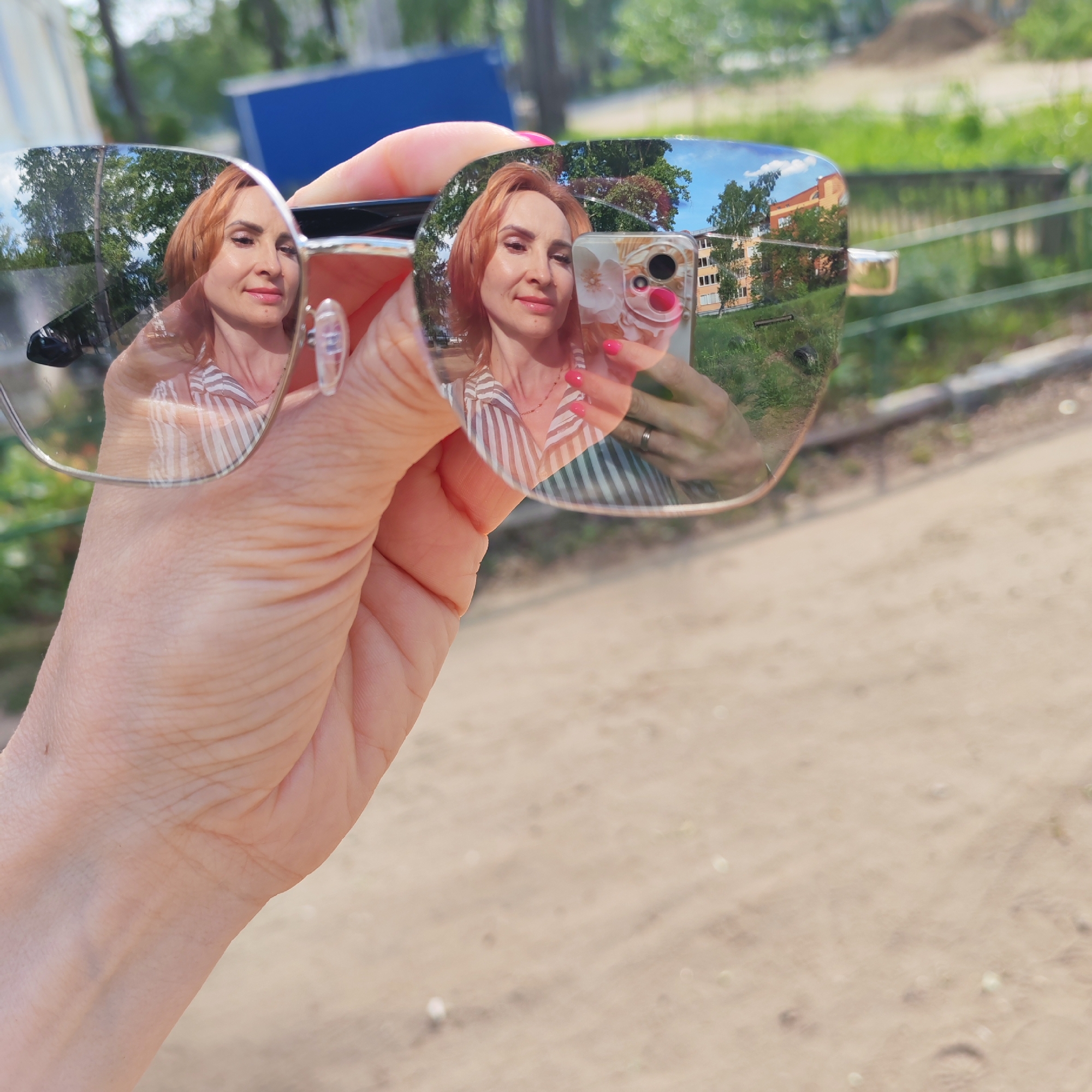 A woman with short, reddish-brown hair is holding a pair of sunglasses in her hand, reflecting her own image. She is taking a selfie with a phone that has a white floral case and pink accents. The reflection in the glasses shows a building, trees, and grass, suggesting an outdoor setting. The background behind the hand and sunglasses appears to be a dirt or gravel surface. The location in the city of Chaikovsky, Russia, can be inferred from details in the reflected scene, such as the building architecture and surrounding vegetation.