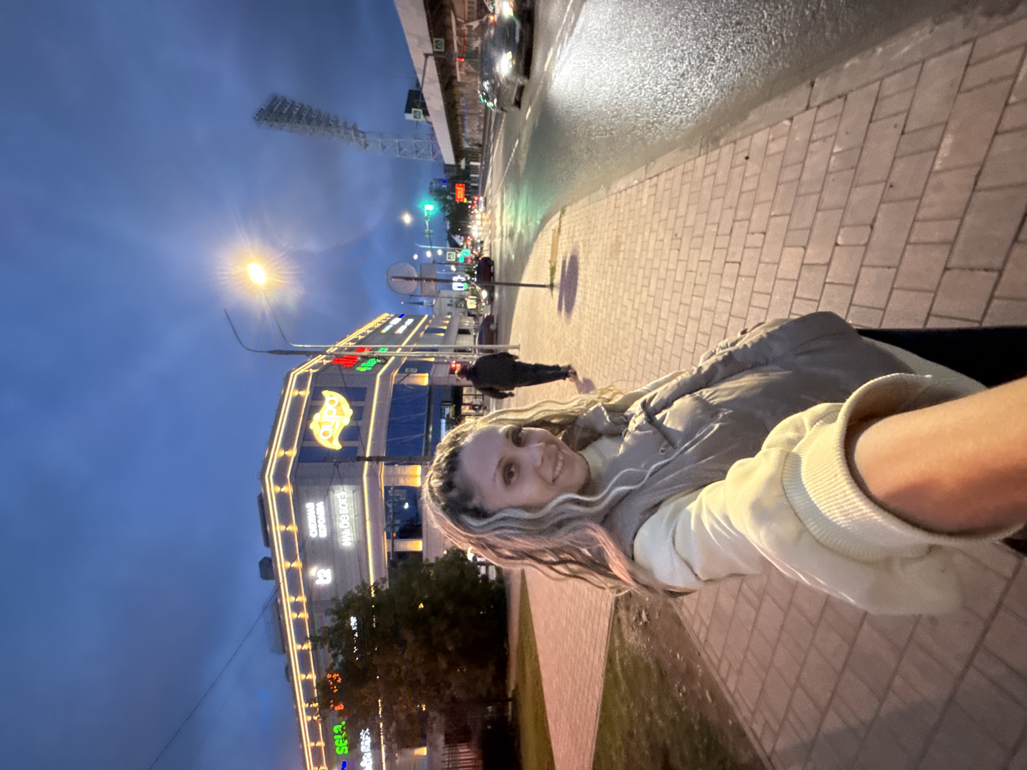 A woman with blonde braided hair and a grey puffer vest over a cream sweatshirt walks towards the camera on a paved sidewalk in a city at dusk. She is smiling. Behind her, a brightly lit building with neon signs is visible. A street with cars and traffic lights extends into the background, with a stadium light tower in the far distance. The sky is dark blue with clouds. The scene appears to be in the city of Rusanovo, Russia, though this cannot be definitively confirmed from the image alone.
