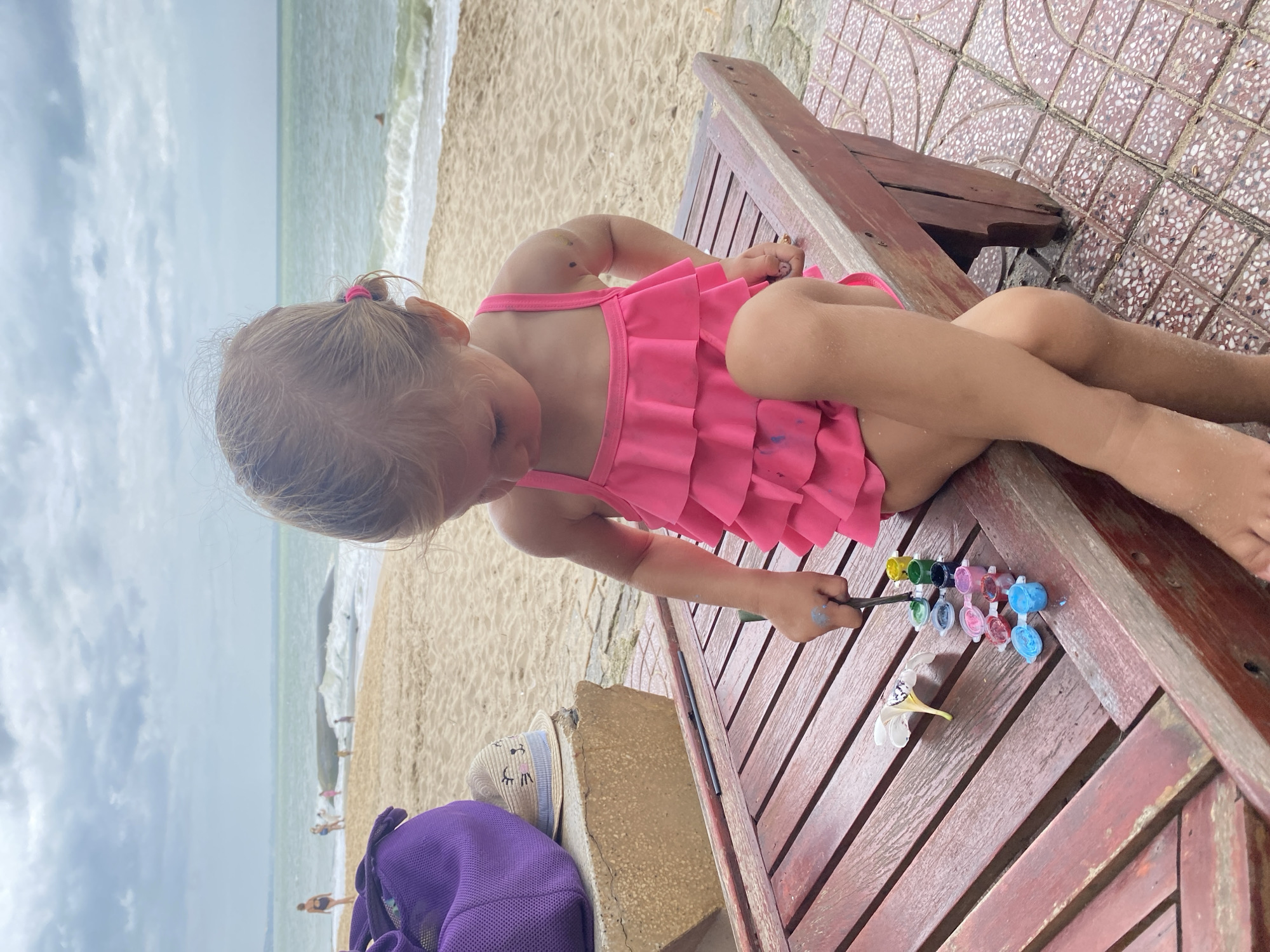 A young girl, with blonde hair in a ponytail, wears a pink ruffled swimsuit and sits on a wooden bench on a beach. She is focused on painting with a brush and small pots of paint spread out on the bench before her. To her left, a white flower lies on the bench, and further back, a purple bag and a straw hat with a cat design are visible on a stone structure. The background reveals a sandy beach with people walking and swimming in the ocean, under a cloudy sky. The location is identified as Mũi Né, Vietnam.