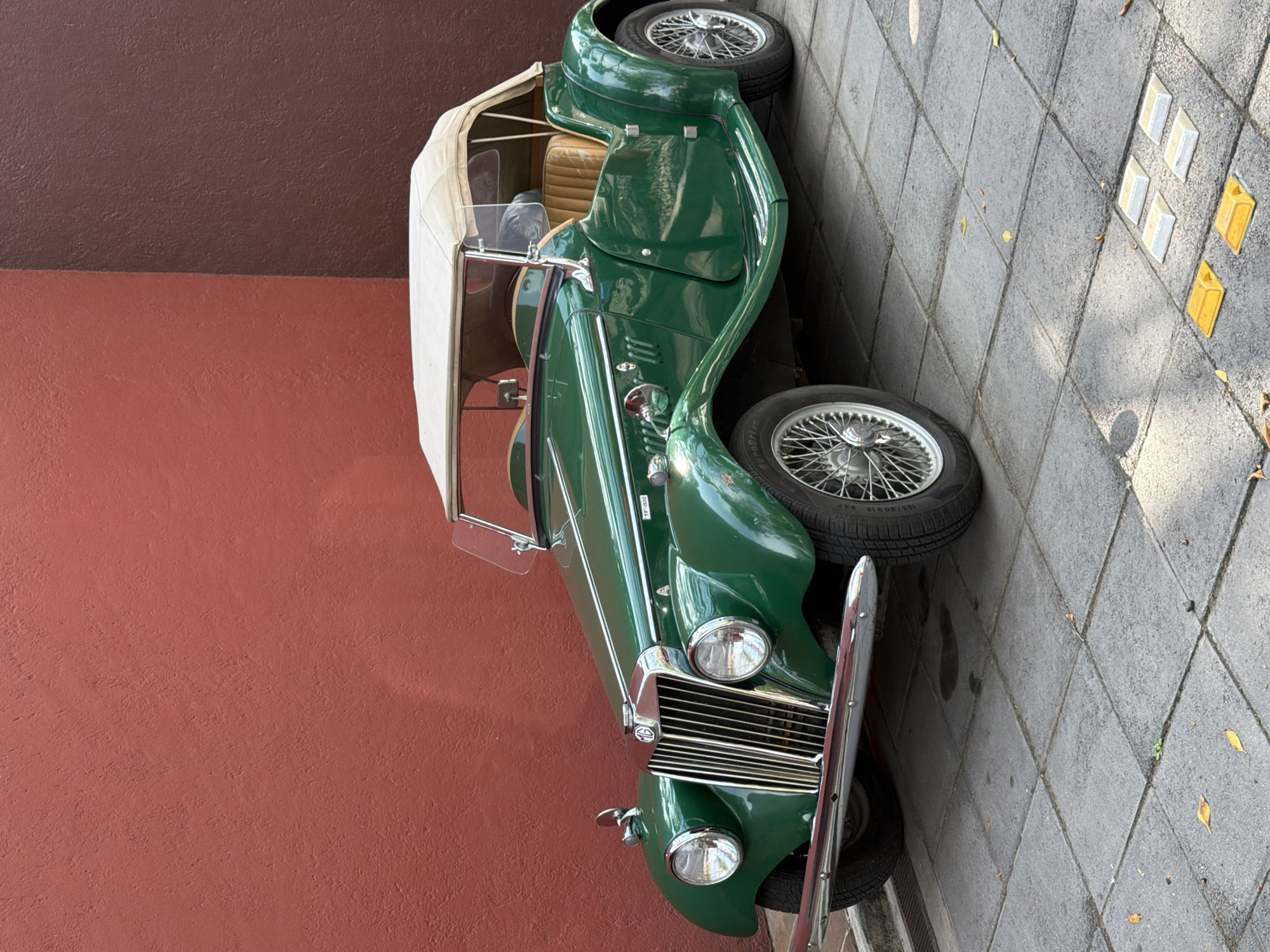 A classic, vintage green convertible car is prominently featured, parked outdoors on a sunny day. The car has a gleaming dark green finish, complemented by numerous chrome accents including a vertical-slat front grille, front bumper, round headlights, side mirrors, and windshield frame. It is equipped with spoked wire wheels and black tires. A cream or off-white soft top is partially visible, appearing to be folded down or retracted, revealing a tan-colored interior with visible seats.
The car is positioned alongside a textured wall, painted in a deep reddish-brown hue. On the other side of the vehicle is a gray sidewalk composed of square tiles, with a few scattered dry leaves. Towards the right side of the image, four small, rectangular road markers are visible on the sidewalk: two white and two yellow. The bright lighting suggests it is daytime, casting shadows on the sidewalk. No people are present in the scene. A small white sticker on the car's side, near the windshield base, displays the text "TF-1030". The setting appears to be an urban or semi-urban area in Puebla, Mexico, with the car simply parked.