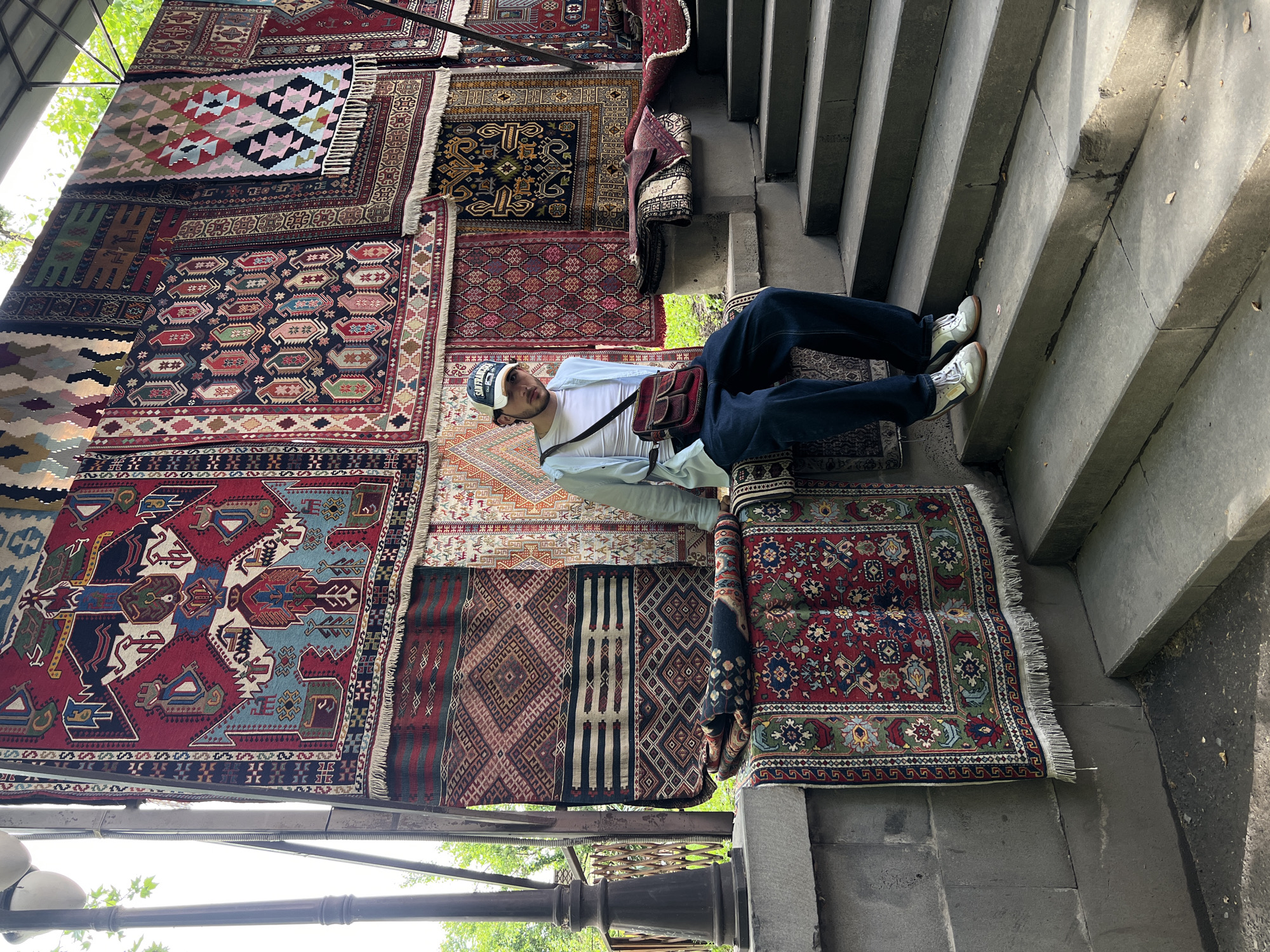 A man is lying on a rug in Yerevan, Armenia. He is wearing a white t-shirt, light blue button-up shirt, jeans, sneakers, and a baseball cap. He has a brown bag slung over his shoulder. He is lying on a rug that is on the ground next to a set of concrete stairs. Behind him is a wall of rugs hanging from a structure. The rugs are of various colors and patterns. The day appears sunny.