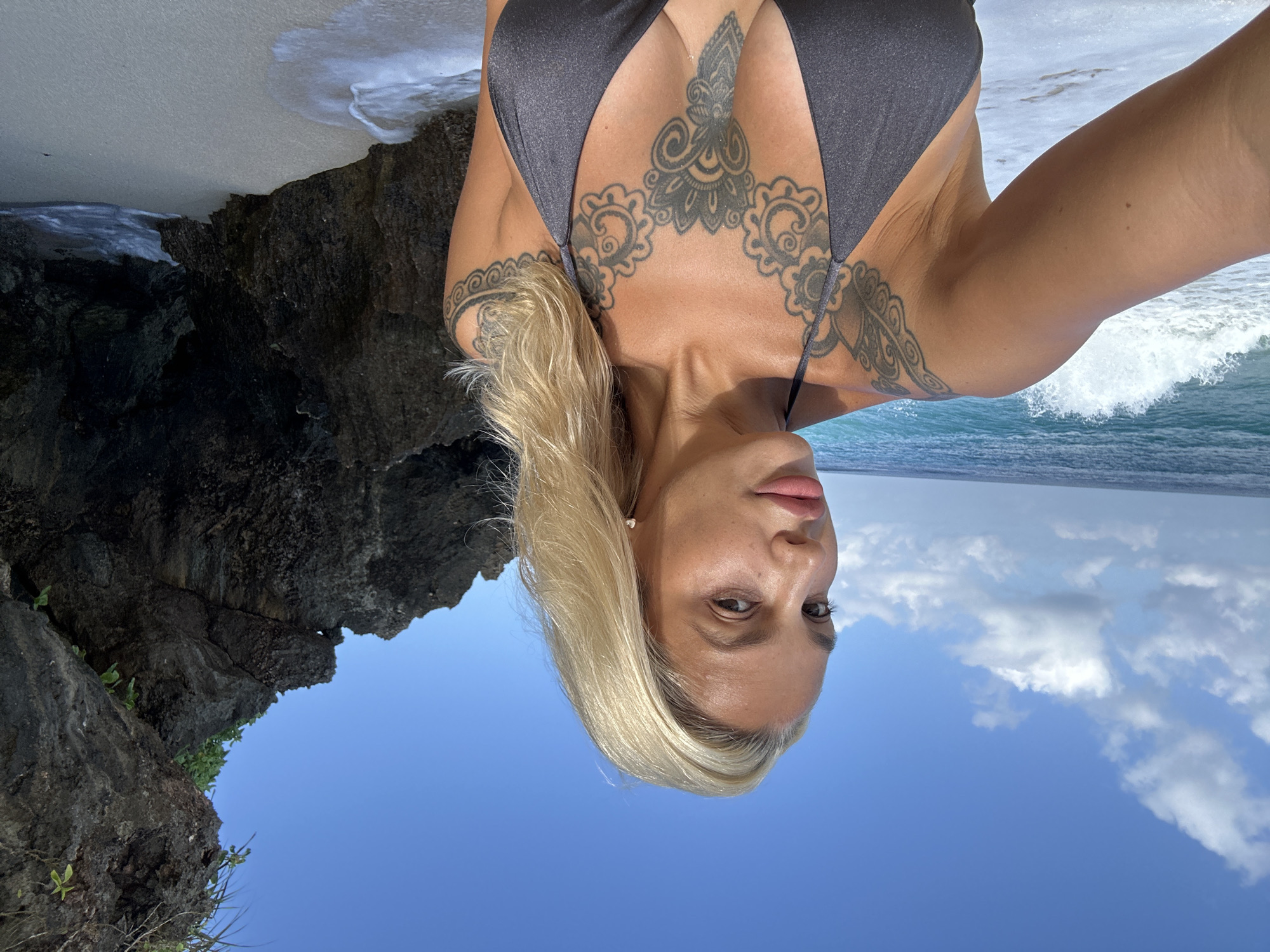 A blonde woman with extensive black tattoos on her chest and left arm stands on a sandy beach in Kuta Selatan, Indonesia. She is wearing a dark grey bikini top and is looking towards the camera. The ocean is visible behind her with waves breaking on the shore. To her right, a large dark rock formation juts out into the scene. The sky is bright blue with scattered white clouds.