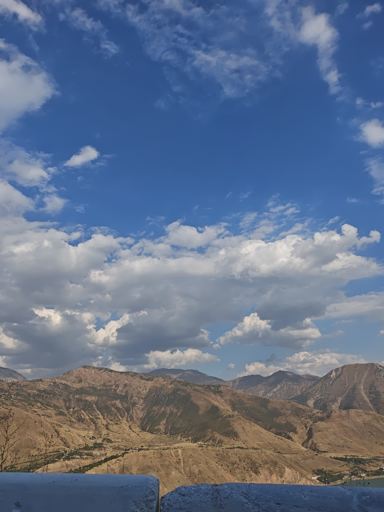 A panoramic view of the mountainous landscape near the city of Yangikurgan, Uzbekistan, is captured under a bright blue sky dotted with white clouds. The foreground features a weathered white concrete barrier, partially obscuring the view of a valley below. The valley floor is a patchwork of dry, golden grasses and scattered green vegetation, with a small cluster of buildings indicating a settlement. Further back, the terrain rises into rugged, brown and tan mountains, characterized by steep slopes, ravines, and sparse vegetation. The sunlight casts shadows, highlighting the contours and textures of the mountains. In the distance, additional mountain ranges fade into a hazy blue, suggesting a vast, expansive natural environment. The overall impression is one of a dry, arid, and remote mountainous region.
