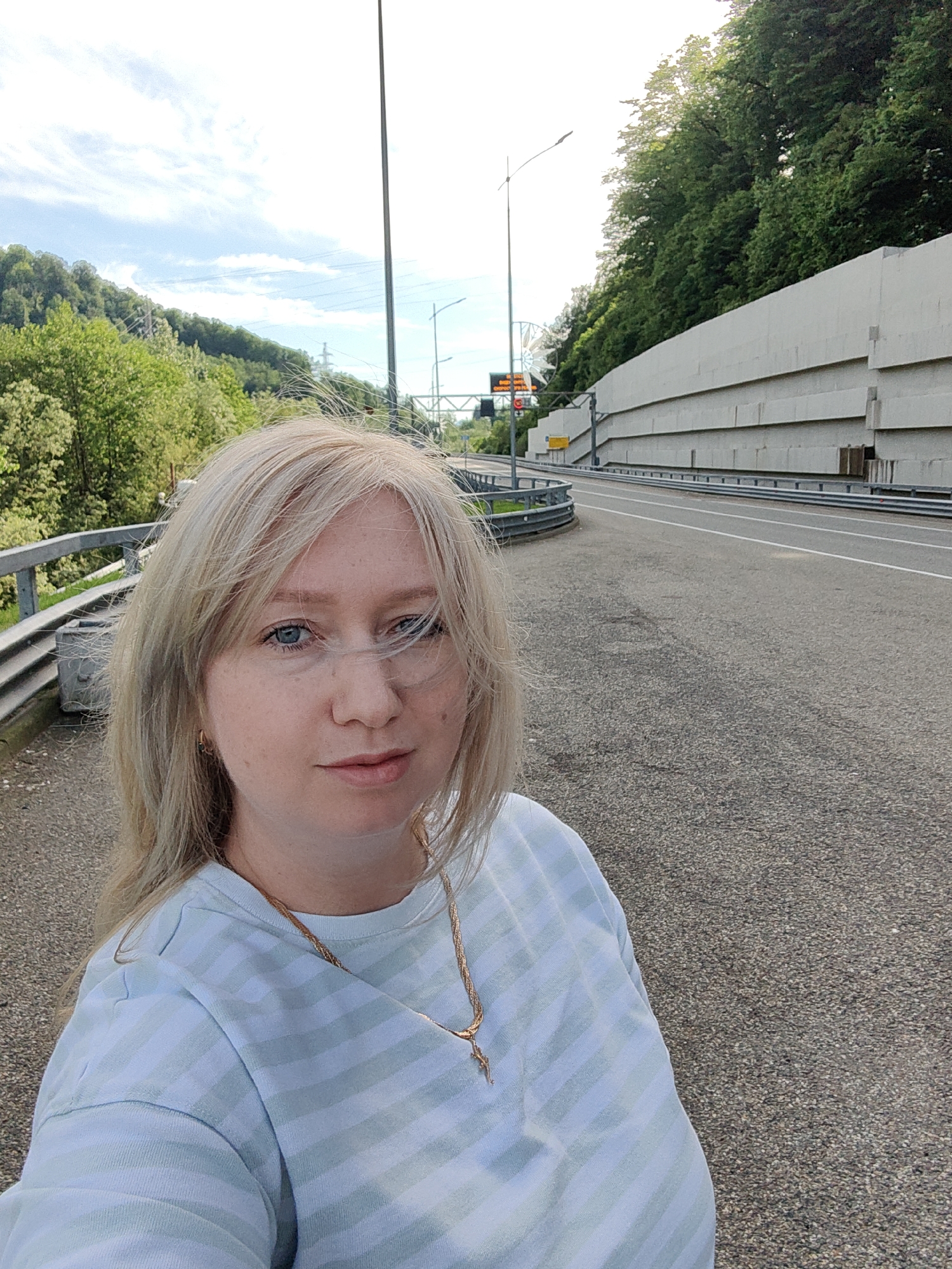 A woman with blonde, windswept hair and blue eyes is captured in a selfie during daytime, looking directly at the camera with a neutral expression. She is wearing a light blue and white striped long-sleeved shirt and a gold chain necklace.
The background reveals a mountainous road environment in Esto-Sadok, Russia. On the left, a steep hillside is densely covered with green trees and foliage, with power lines visible against a partly cloudy sky. On the right, a multi-tiered concrete retaining wall supports another forested slope, bordering an asphalt road that curves to the right. The road is lined with metal guardrails.
Further along the road, a streetlight, a red traffic light, and a small yellow road sign are visible. A blurred white electronic sign displays Russian text, partially discernible as "ВНИМАНИЕ" (Attention), "ВРЕМЕННО" (Temporarily), "ОГРАНИЧЕНО" (Limited), and "ДВИЖЕНИЕ" (Traffic/Movement), indicating a temporary traffic restriction. The weather is bright with diffused sunlight, suggesting a clear to partly cloudy day.