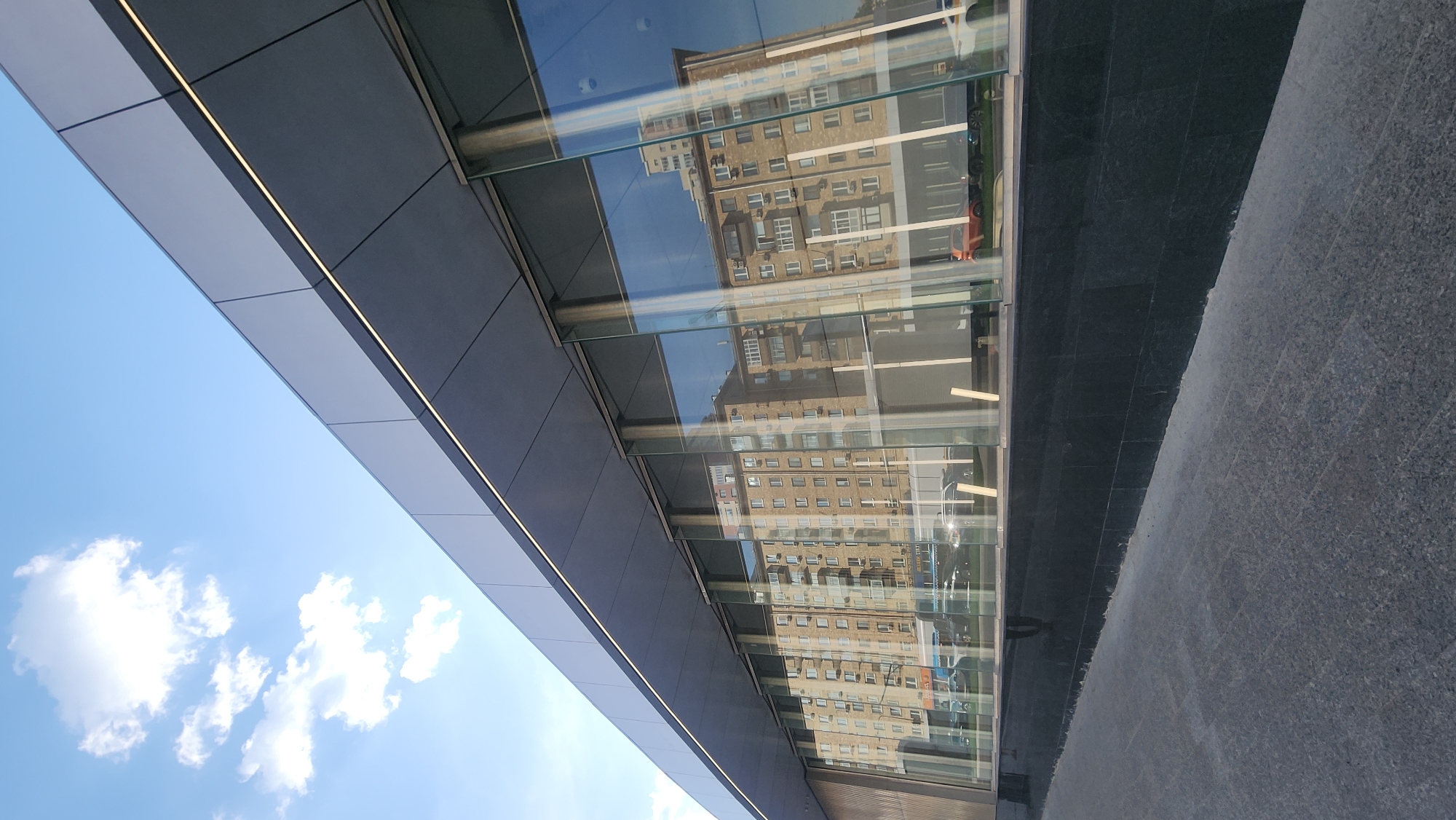 The image captures an architectural view, likely in an urban environment. A modern building with a sleek, angular design is visible, featuring a combination of gray cladding and large glass panels. The glass surfaces reflect the surrounding cityscape, showing an older building with a facade of multiple windows. The sky, dotted with fluffy white clouds, is reflected as well. The ground appears to be paved with dark-colored stone slabs.
Based on the architecture and reflections, the location is likely in the city of Moscow, Russia.