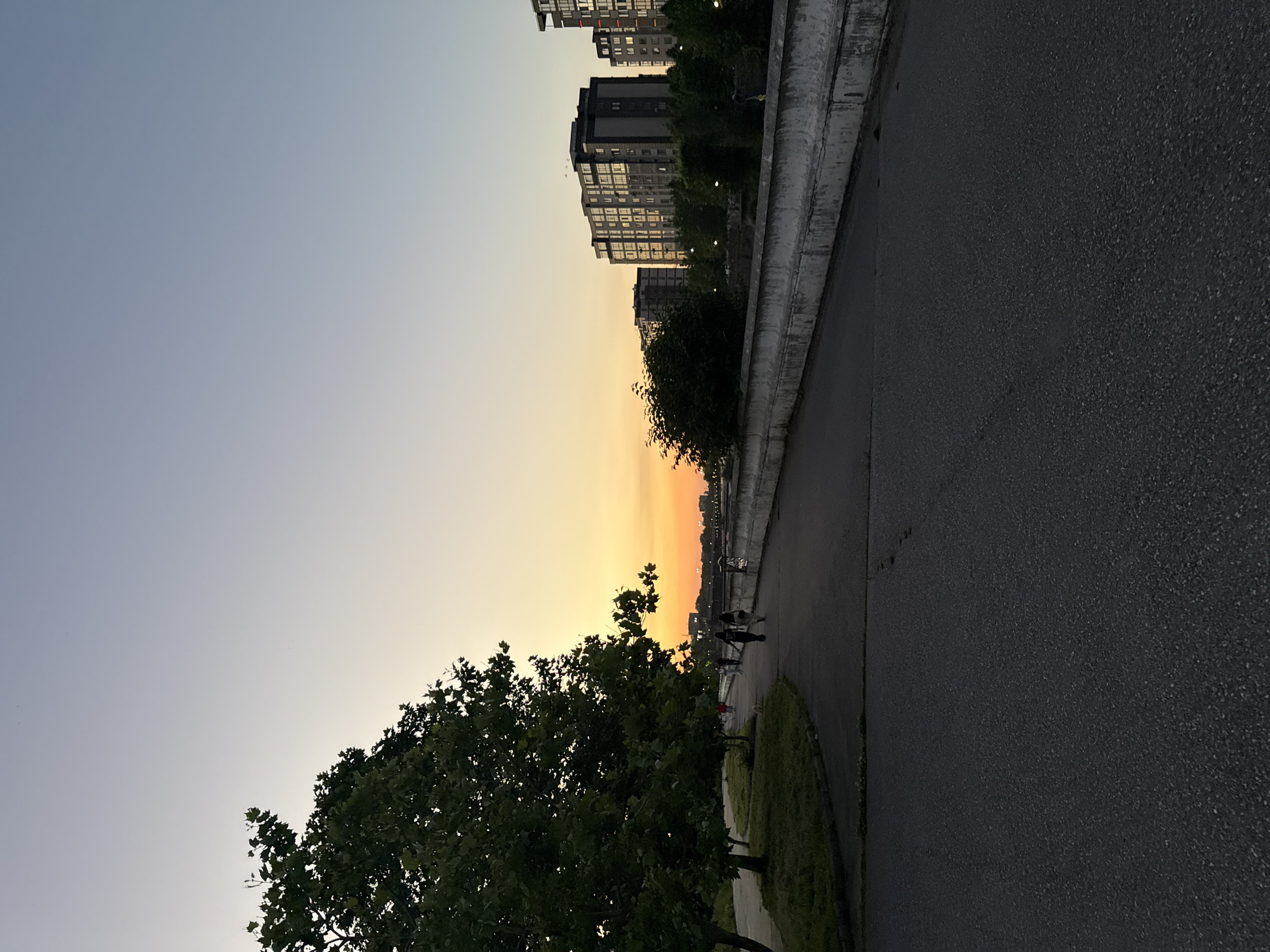 The image depicts a sunset scene along a paved walkway in Khachta, Russia. A large tree with dense green foliage is situated on the left side of the walkway, with grass and smaller trees behind it. Several people are visible walking along the path, some appearing to be couples. On the right side of the walkway, a concrete barrier separates it from a body of water, beyond which are several tall, multi-story apartment buildings. The sky transitions from a pale blue at the top to a vibrant orange and yellow near the horizon, indicating the setting sun. The overall atmosphere is calm and serene, suggesting an evening stroll.