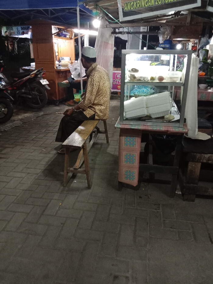 The scene depicts a nighttime outdoor market or street food area in Turunsih, Indonesia, illuminated by artificial lights. The ground is paved with interlocking grey concrete blocks.

An adult male is seated on a simple wooden bench, facing right and slightly away from the camera. He wears a white skullcap (peci), a long-sleeved light-brown patterned shirt, and dark trousers. His right hand holds a thin, lit cylindrical object consistent with a cigarette, positioned near his lap.

To the right of the seated male, a metal-framed glass display case rests on a table draped with a pink and blue patterned fabric skirt. The case contains various food items on its two shelves, with a substantial stack of white, rectangular disposable containers on the lower shelf. An overhead banner partially displays the text "ANGKRINGAN".

In the background to the left, under a blue tarp canopy and prominent artificial lighting, a wooden food stall is visible. A second individual is partially seen, seated behind this stall, wearing dark clothing and green flip-flops. A dark-colored motorcycle is parked immediately to the left of the food stall. Signage on the left stall includes the word "DONAT". The environment features temporary structures and overhead wiring.