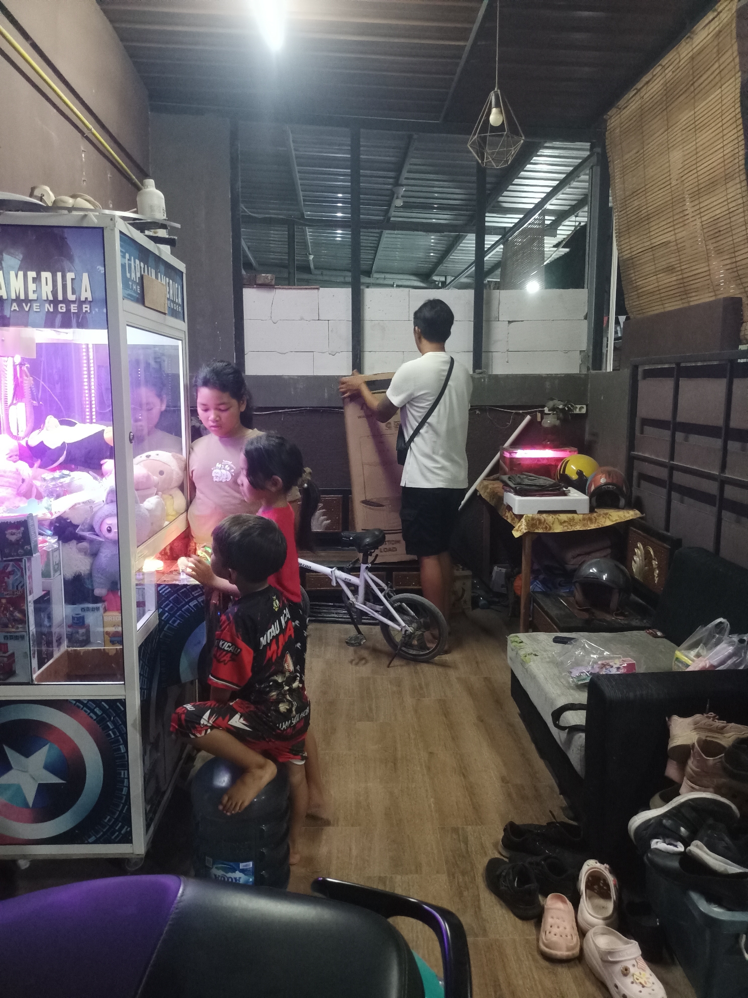 In an indoor setting resembling a shop or storage area in Mranggen, Indonesia, a claw machine adorned with Captain America imagery stands prominently on the left. Inside the machine, various plush toys are visible. A group of individuals, including at least three children and one adult woman, are gathered around the claw machine, seemingly engaged in playing or observing. One child is seated on a large water dispenser behind the machine. In the background, a man is seen holding a large cardboard box. The environment includes a metal roof structure, shelves with items, and a table with helmets and a red glowing object. A small white bicycle is also present. The floor is wooden. Various footwear is scattered near a couch on the right side of the image. The lighting suggests it is either evening or an indoor setting with artificial illumination.