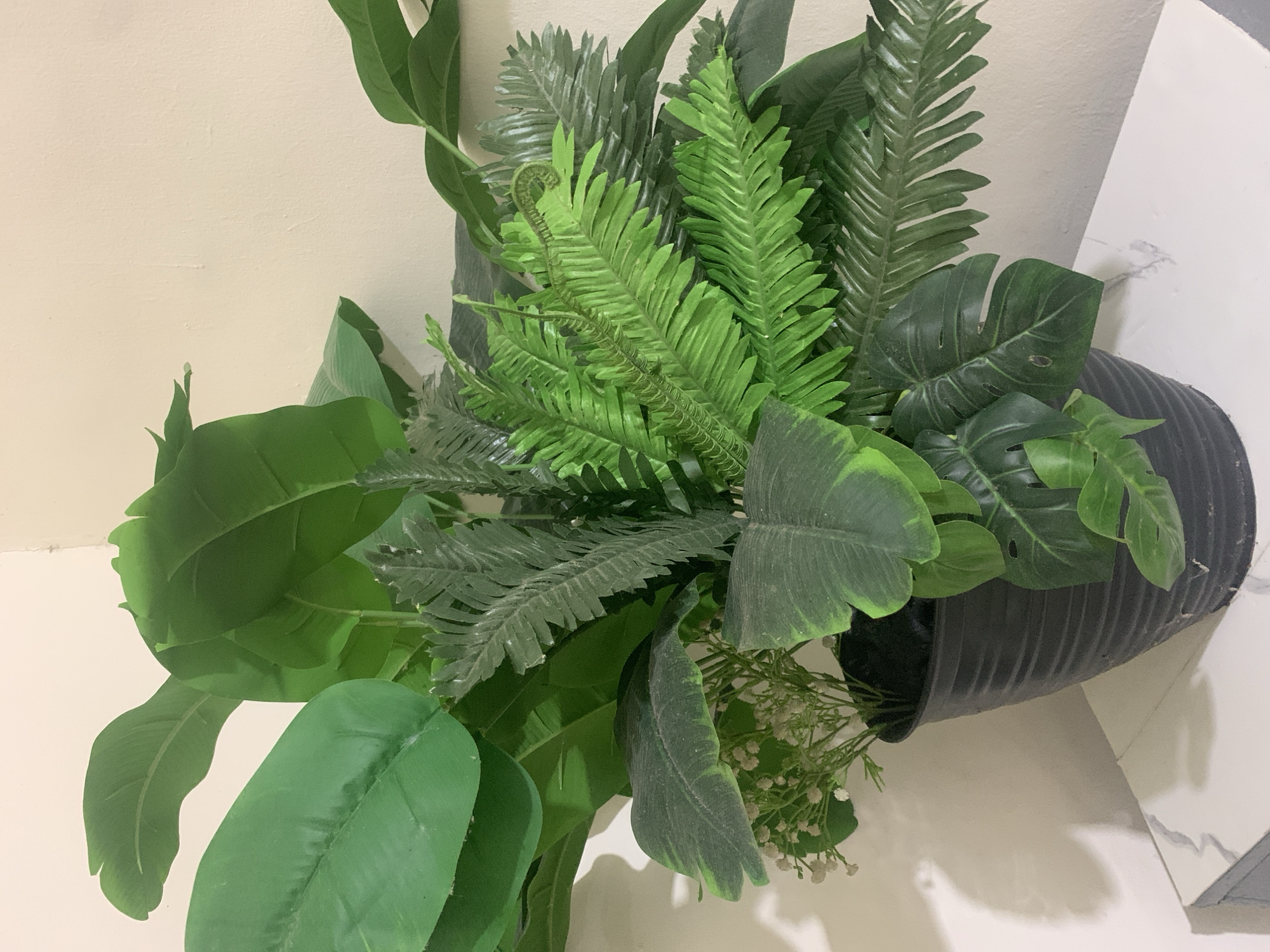 A potted artificial plant is placed on a white surface against a light-colored wall. The plant consists of various green leaves, including large, broad leaves reminiscent of banana leaves, and frilly fern-like leaves. Some smaller white flowers are interspersed within the foliage. The plant is in a ribbed black plastic pot. The background wall has a subtle shadow pattern cast upon it, suggesting a light source to the left of the scene. The surface the pot rests on has a subtle marbled pattern. There are no people present in the image. The location is not discernible from the image itself; any mention of Kuala Pembuang, Indonesia is external information.