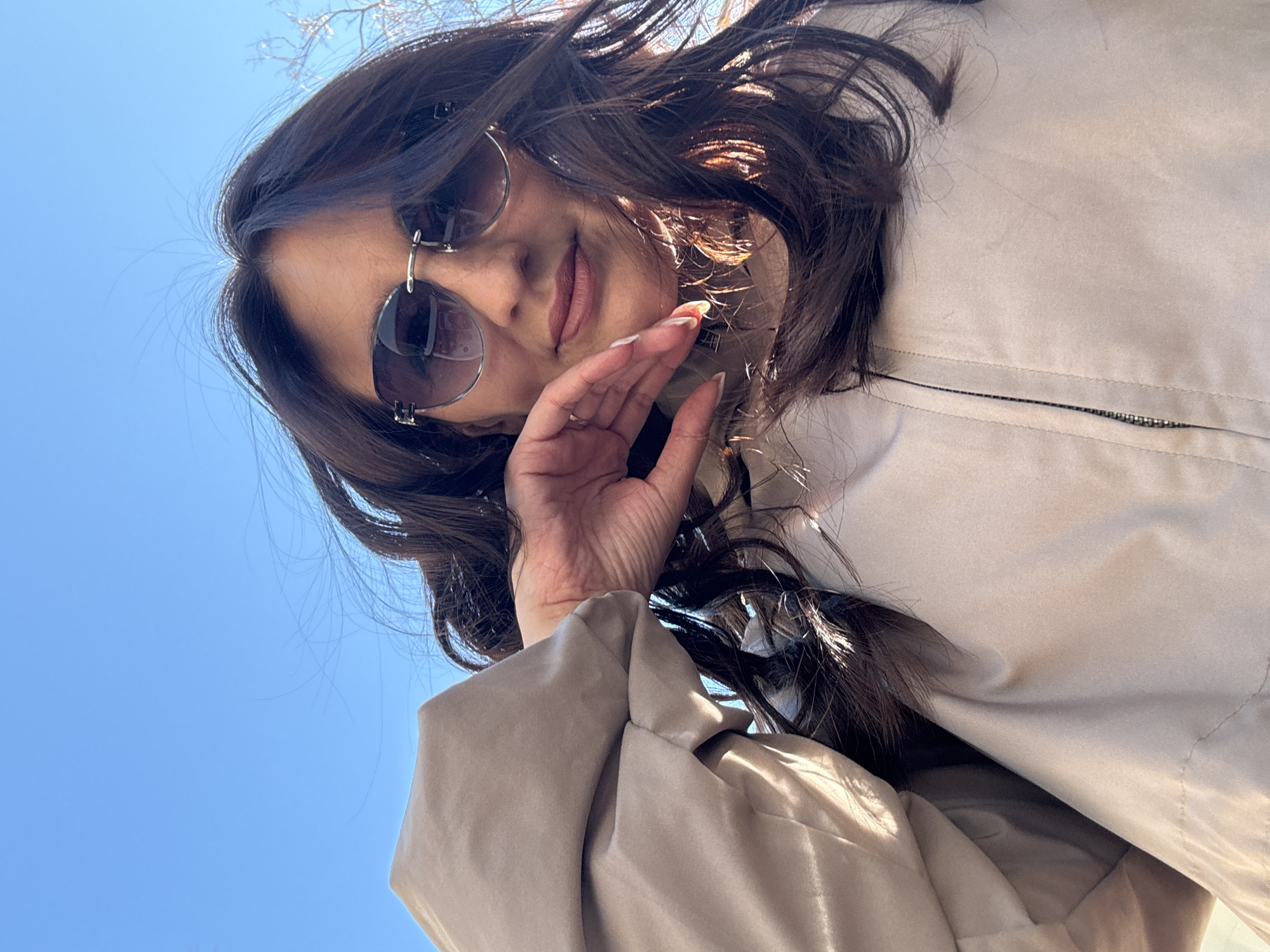 A woman is photographed from a low angle against a clear blue sky. She has dark brown hair, part of which is blowing in the wind, and wears sunglasses with round lenses. She is wearing a light beige jacket with a visible zipper. Her right hand is raised near her face. The specific location in Sochi, Russia, is not evident from the image.