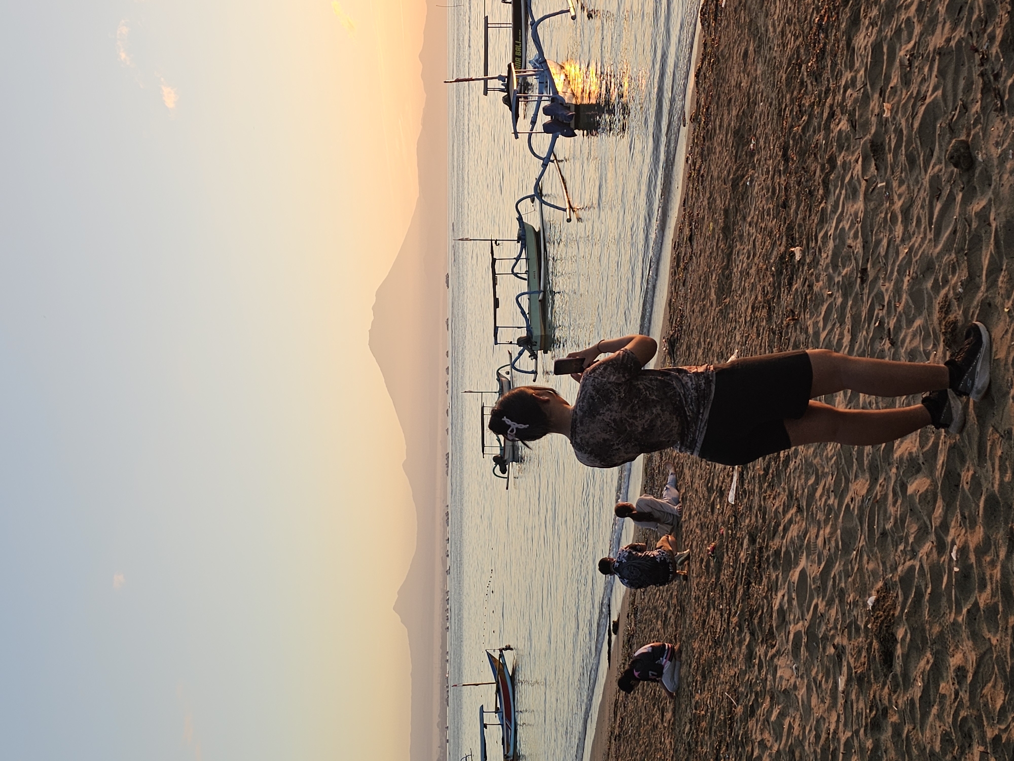 The photograph captures a sunset scene on a beach in Denpasar, Indonesia. The beach has a dark sand texture and the shoreline is lapped by gentle waves. In the background, a mountain silhouette stands against the fading light of the sky, which is a gradient of blues and oranges. Several outrigger boats are visible on the water.
A person, presumably a tourist, is standing on the beach, taking a picture with their phone. Several other people are squatting near the shoreline.